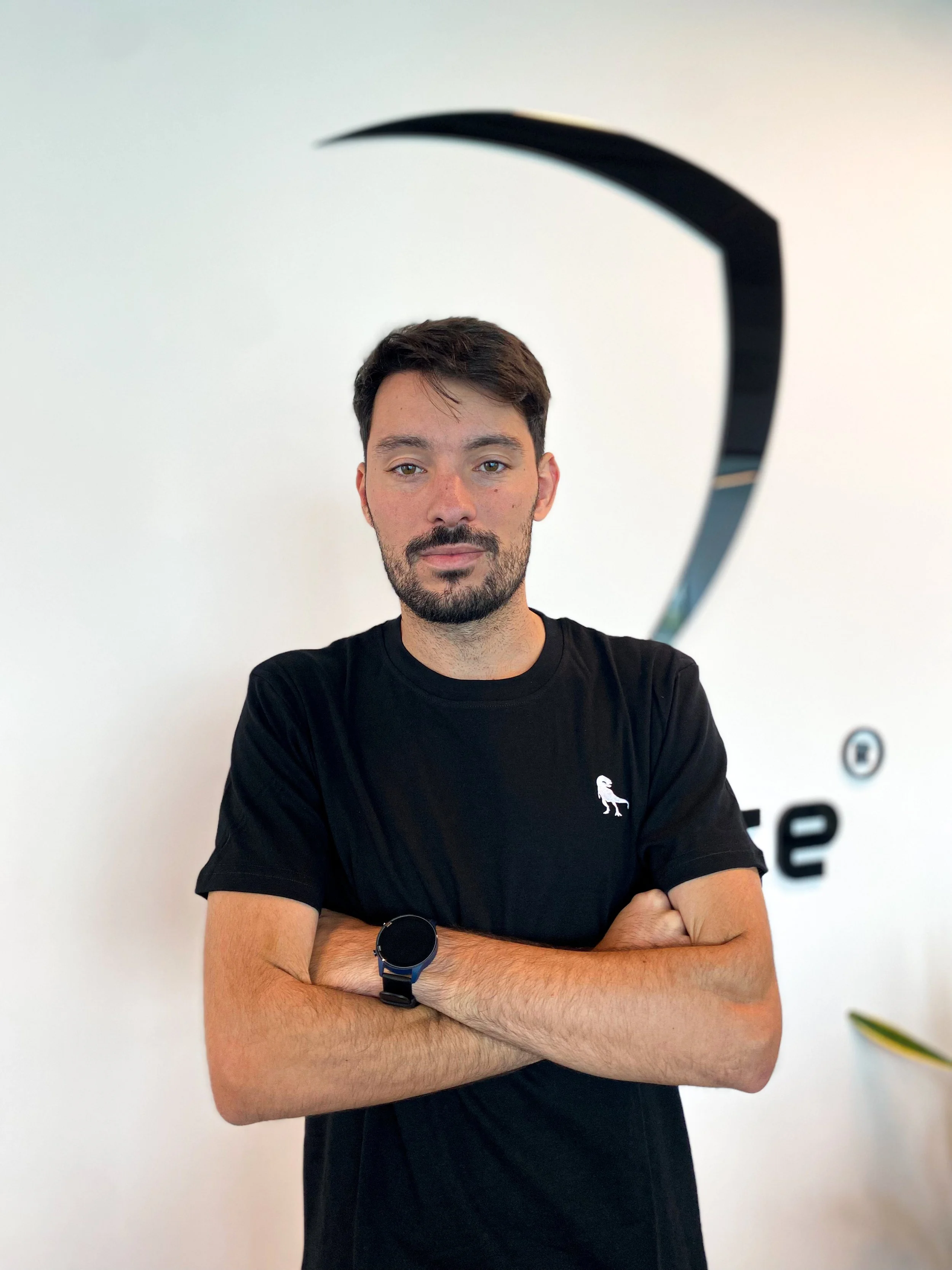 A man wearing a black polo shirt and a black baseball cap with a dinosaur logo stands on a grassy sports field with a scoreboard and trees in the background.