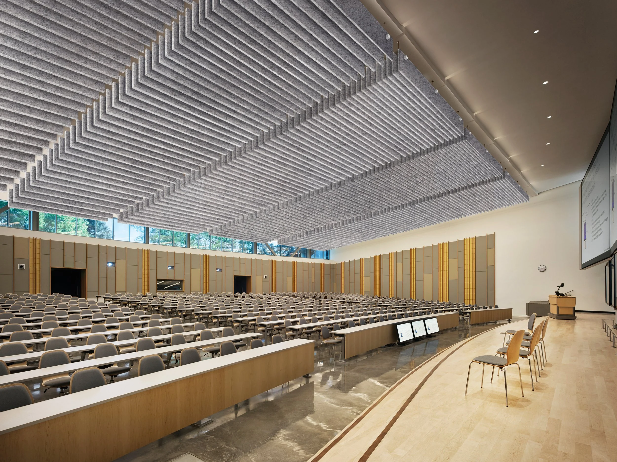 Large conference room with rows of chairs and desks, high ceiling with sound-absorbing GiosFelt Single Baffles hanging from the ceiling, and a large screen at the front.