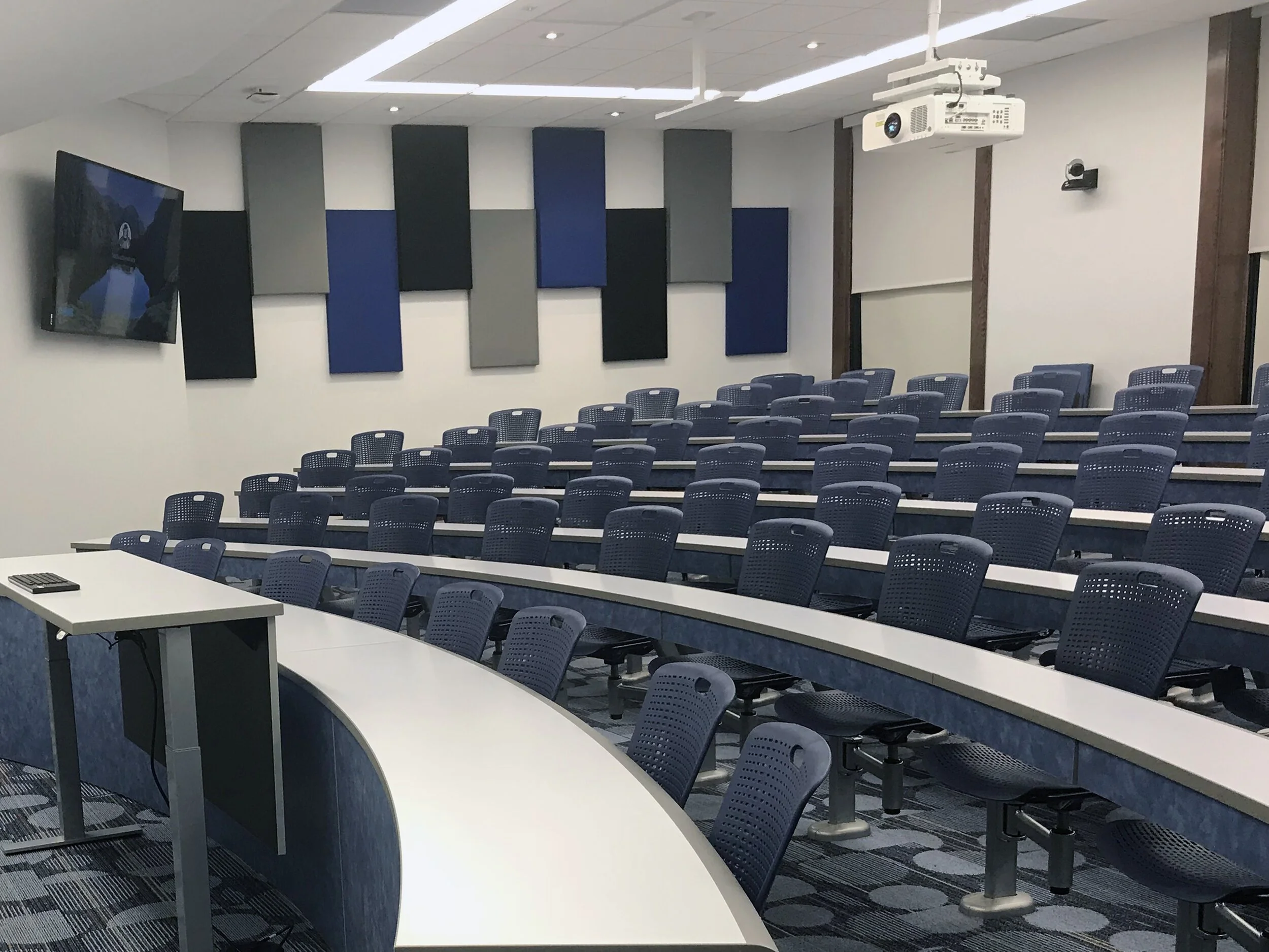 Empty classroom with tiered desks, blue chairs, and a white projector suspended from the ceiling. A large screen is mounted on the front wall, and acoustic Livello L200 Standard wall acoustic panels on the walls.