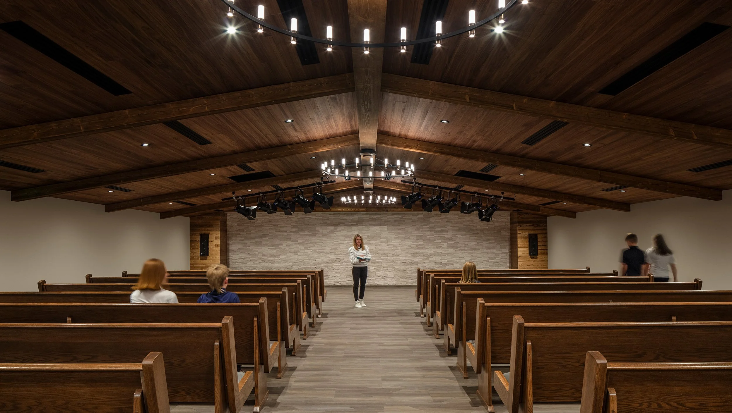 A woman standing on a stage in a large room with WoodLine Parallel acoustical ceiling paneling, wooden ceiling beams, surrounded by empty wooden pews, with a few people seated and engaging in conversation.