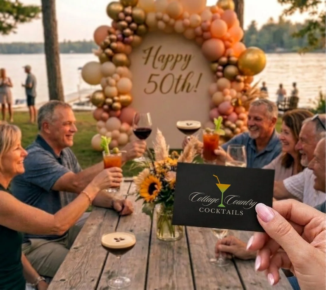 People celebrating an 50th birthday by a lake on a wooden deck, with a large balloon display that says 'Happy 50th!', a table with drinks, a sunflower centerpiece, and a person holding a cocktail card that says 'Cottage Country Cocktails'.