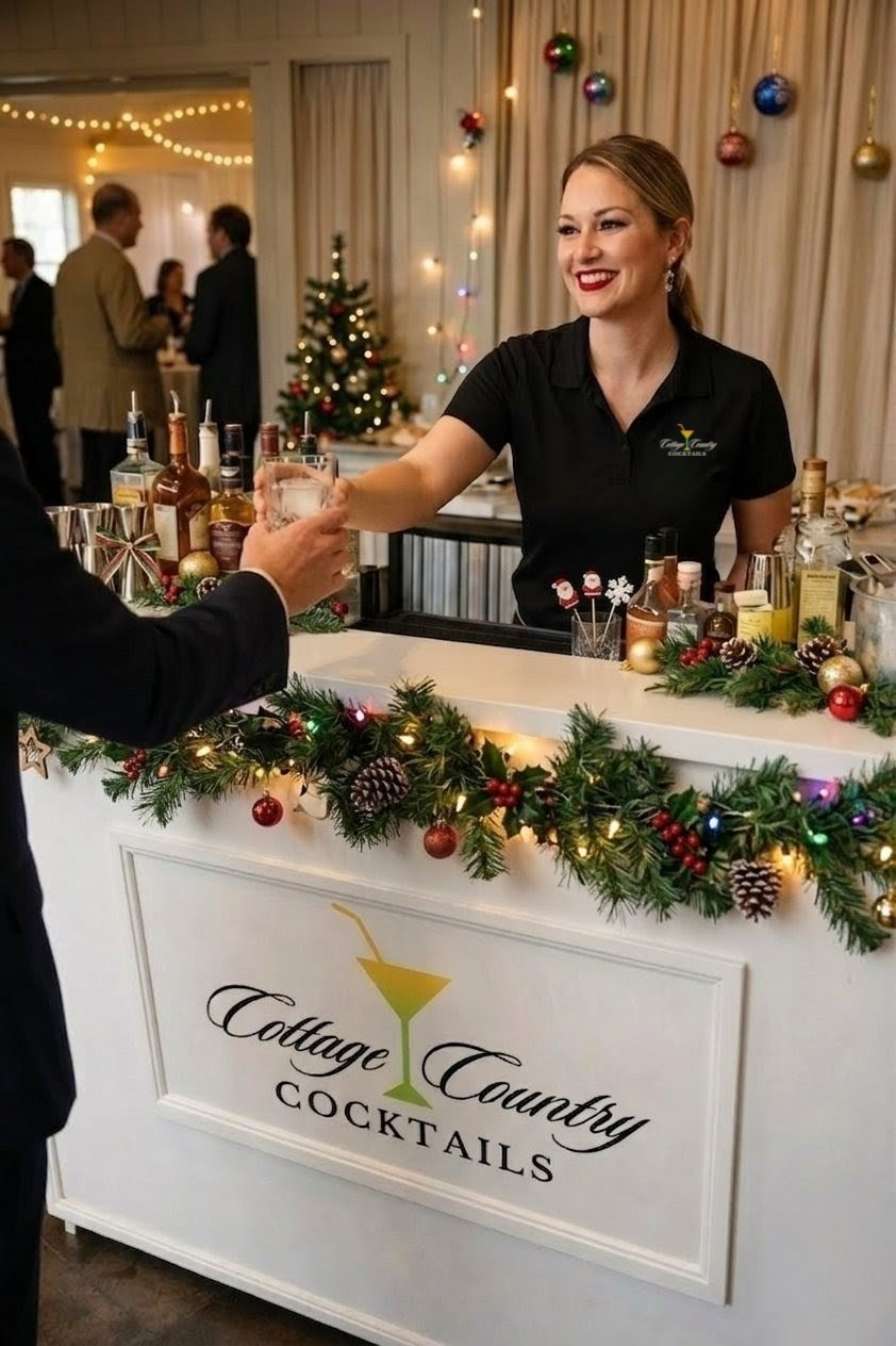 A smiling female bartender wearing a black polo shirt with the Cottage County Cocktails logo, serving a drink to a customer at a holiday-themed bar decorated with Christmas ornaments, pinecones, and garland, with a decorated Christmas tree in the bac