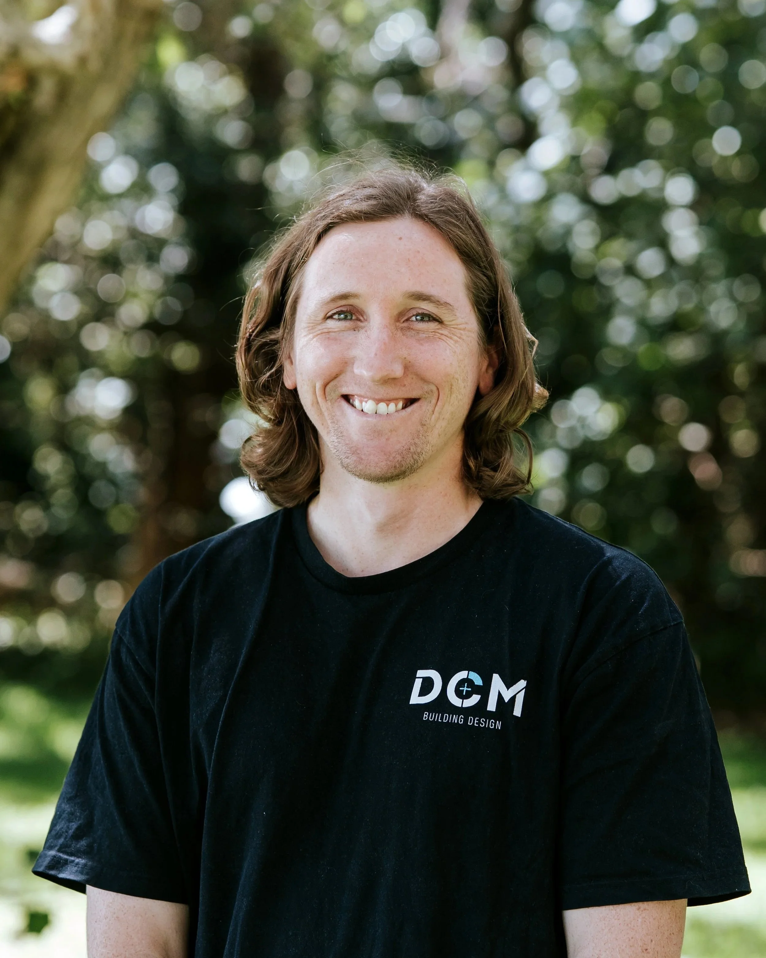 Man wearing a black T-shirt with "DCM Building Design" logo, standing near a body of water with trees in the background.