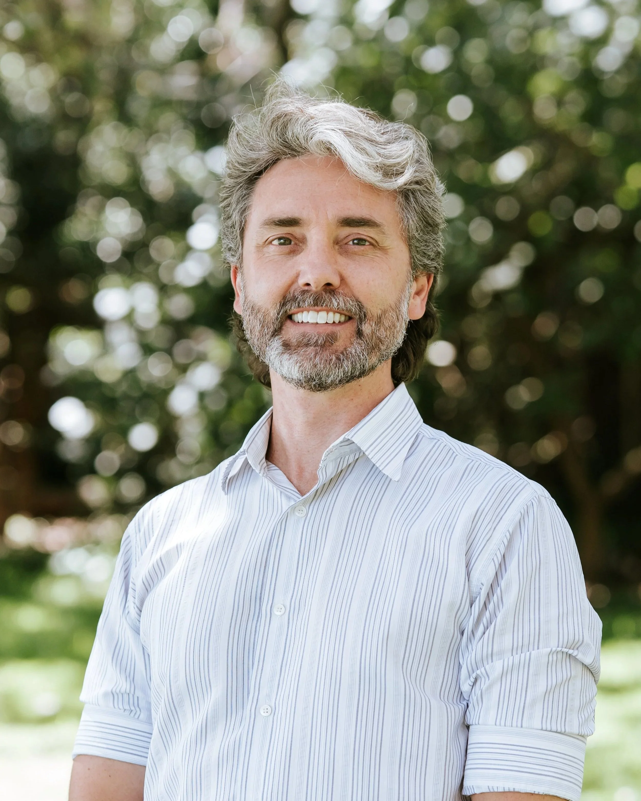 A man with a beard, wearing a black jacket and a striped shirt, standing in front of a building with a modern architectural design featuring vertical wooden slats.