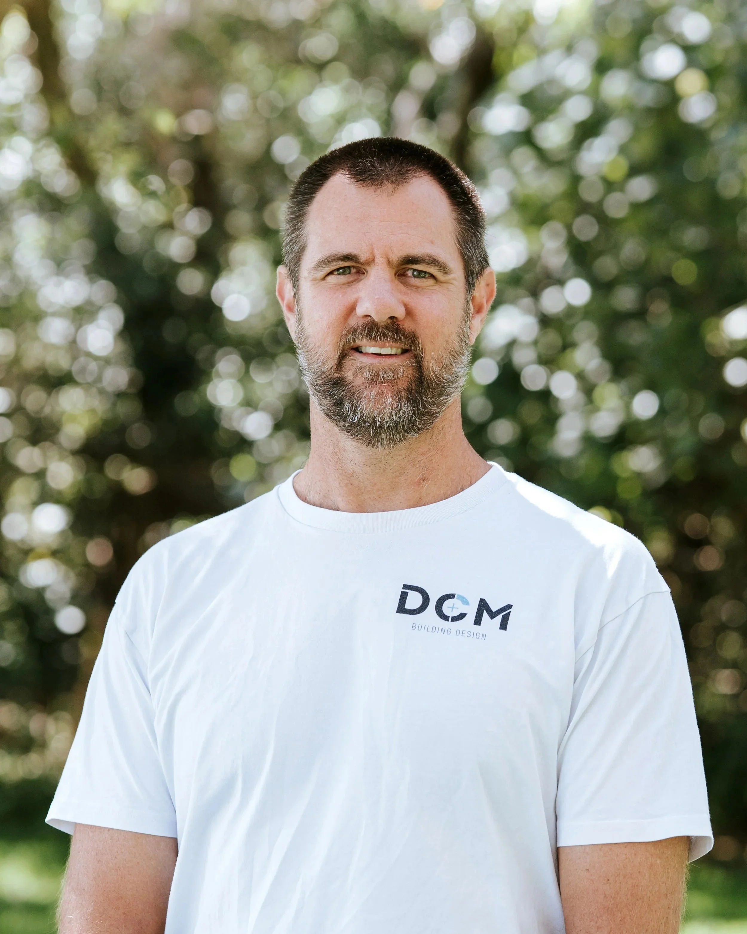 Man with glasses wearing black DCM Building Design t-shirt, standing near a body of water with trees in the background.