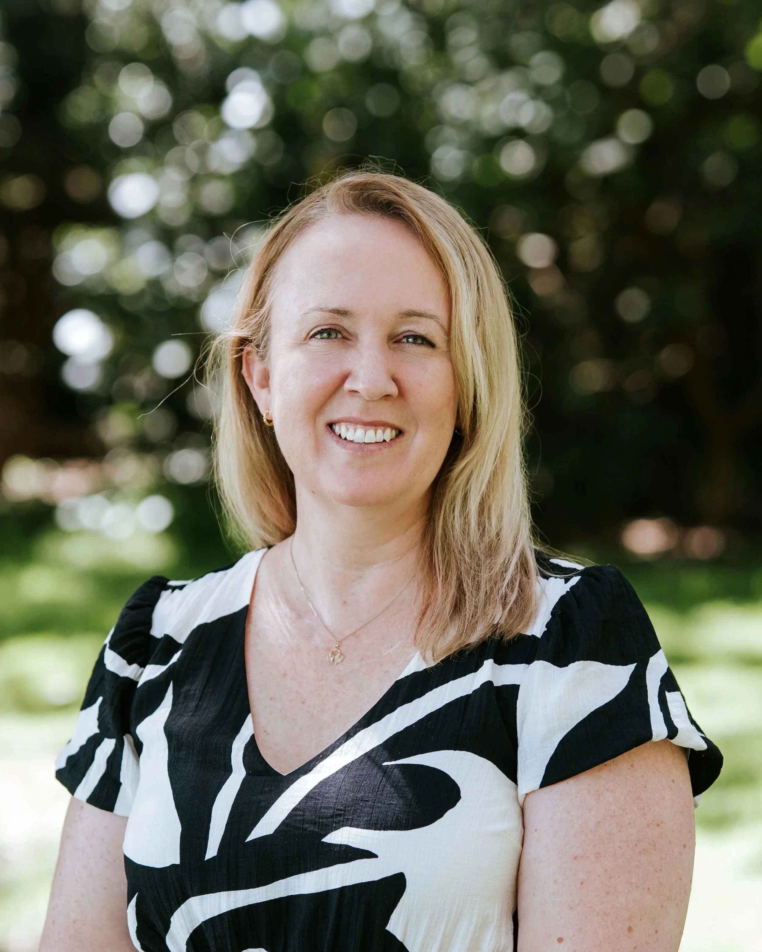 Smiling woman in a black shirt with "DCM Building Design" logo standing outdoors near water with trees in the background.