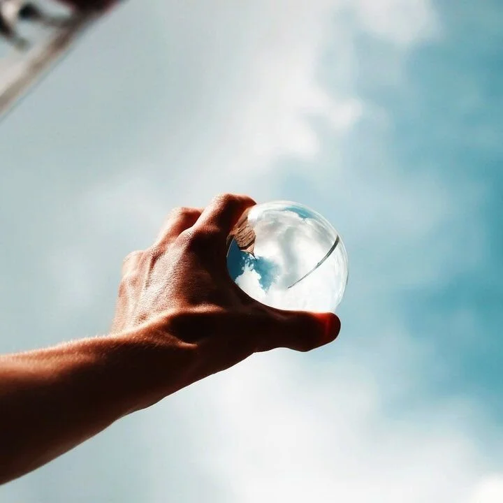 A hand holding a clear glass sphere against a blue sky with clouds, creating a reflection of the sky inside the sphere.