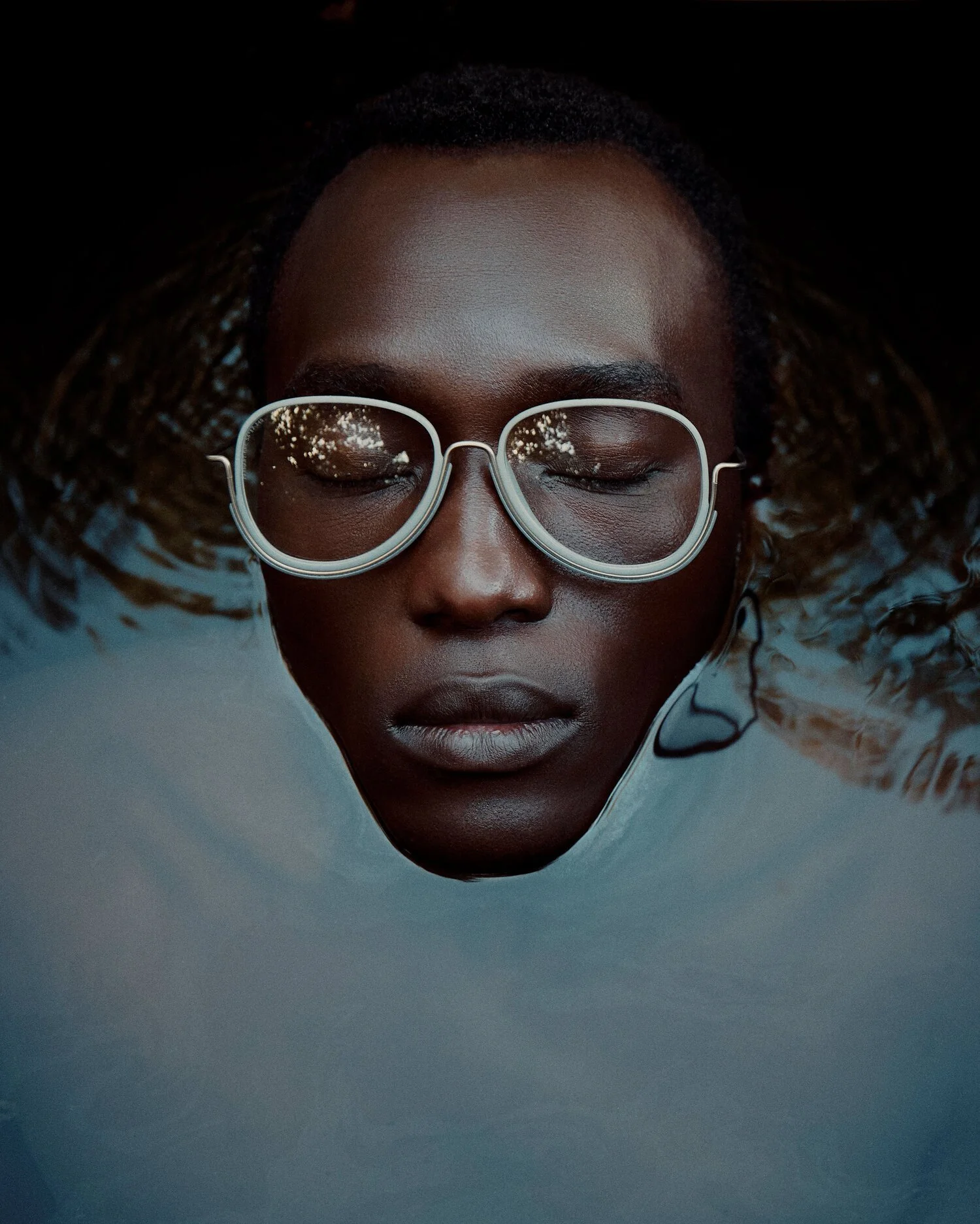 Close-up of a young woman with dark skin and glasses, floating in water with her eyes closed, and her hair spread out around her