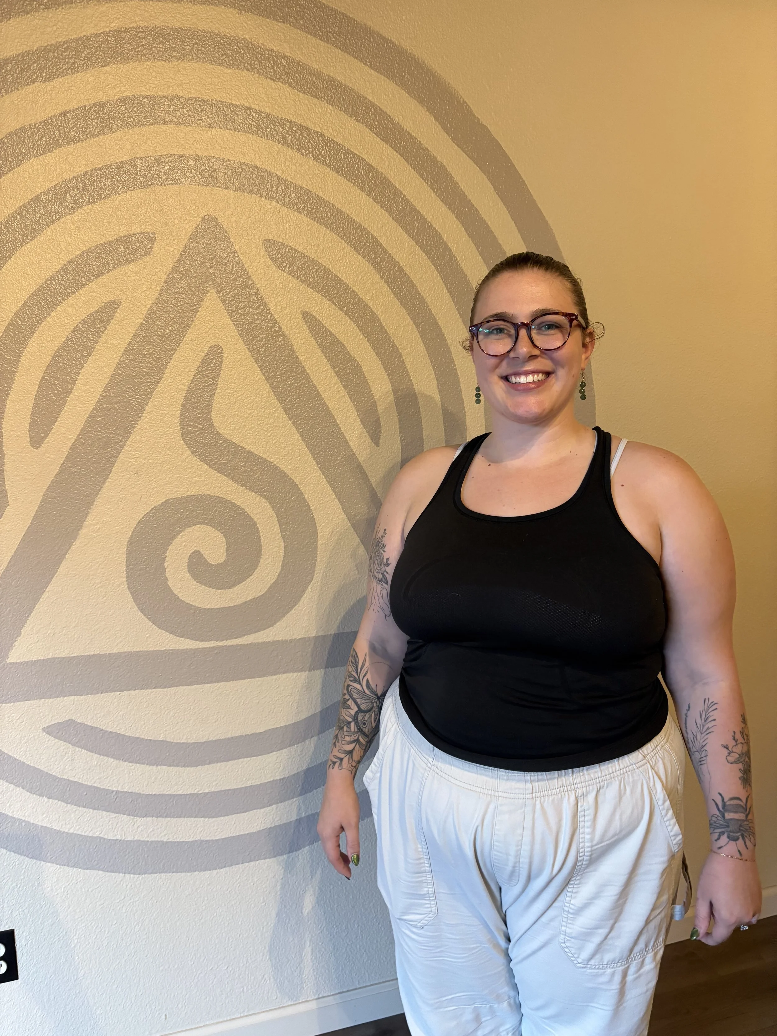 A woman with glasses and earrings smiling, wearing a black tank top and beige pants, standing in front of a wall with a large, decorative, spiral design.