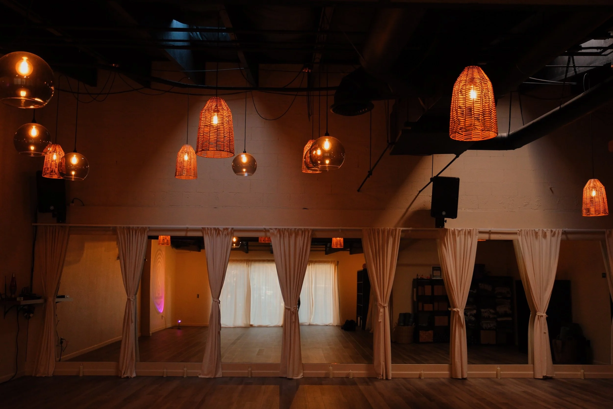 Empty dance studio with wooden floor, cream curtains dividing the space, and warm hanging pendant lights.