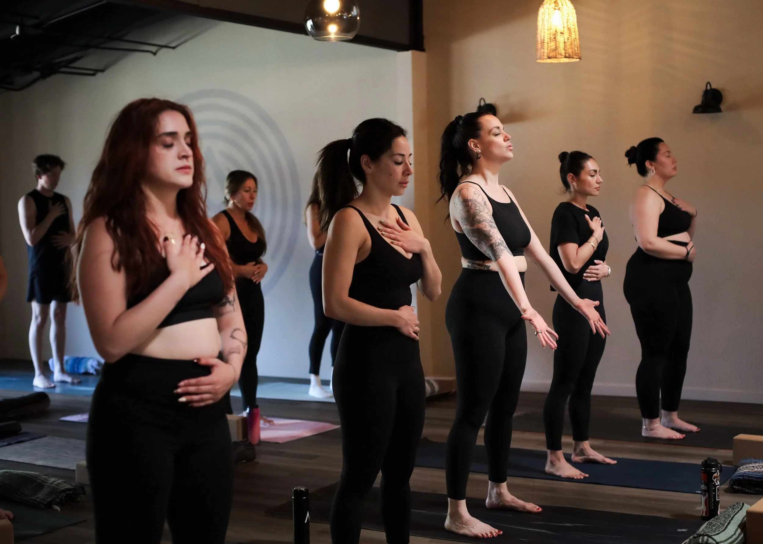 A group of women practicing yoga in a studio, standing with their hands over their hearts and eyes closed, on yoga mats