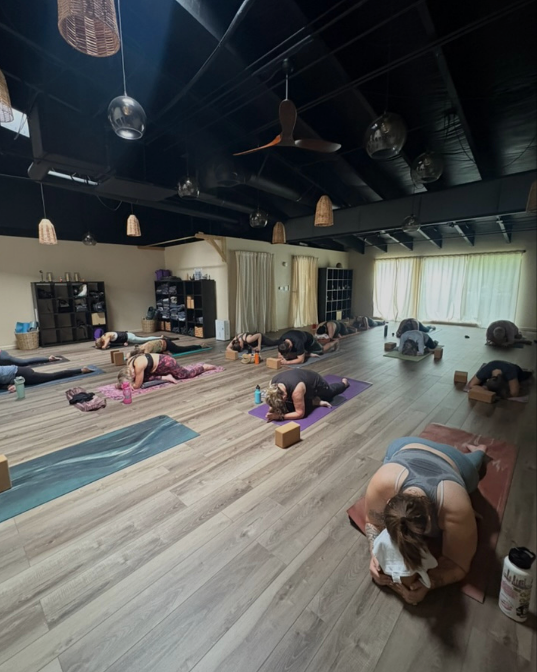 People participating in a yoga class in a spacious studio with wooden floors and natural light, practicing a forward bend pose.