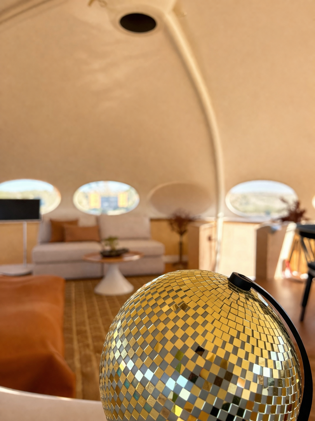 A close-up of a ball-shaped lamp with a mosaic pattern in a living room, featuring a beige sofa, a round coffee table, potted plants, and windows in the background.