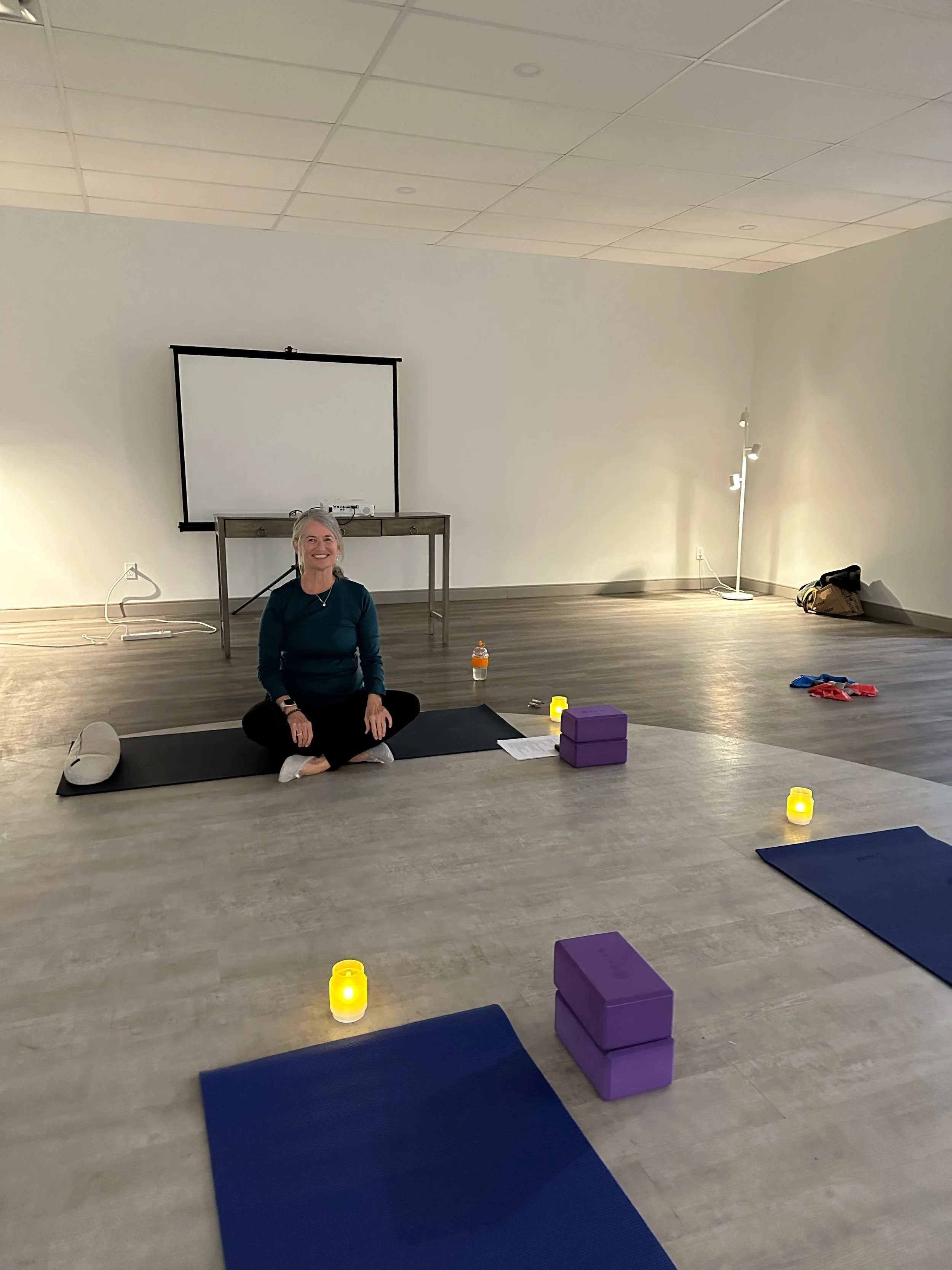 Woman seated cross-legged on a yoga mat in a studio with yoga blocks, candles, and a projection screen.