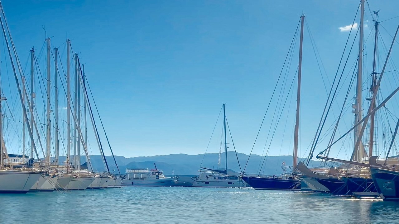 Greek harbor with sailboats, calm blue water, tall masts, distant hills, and soft coastal light.