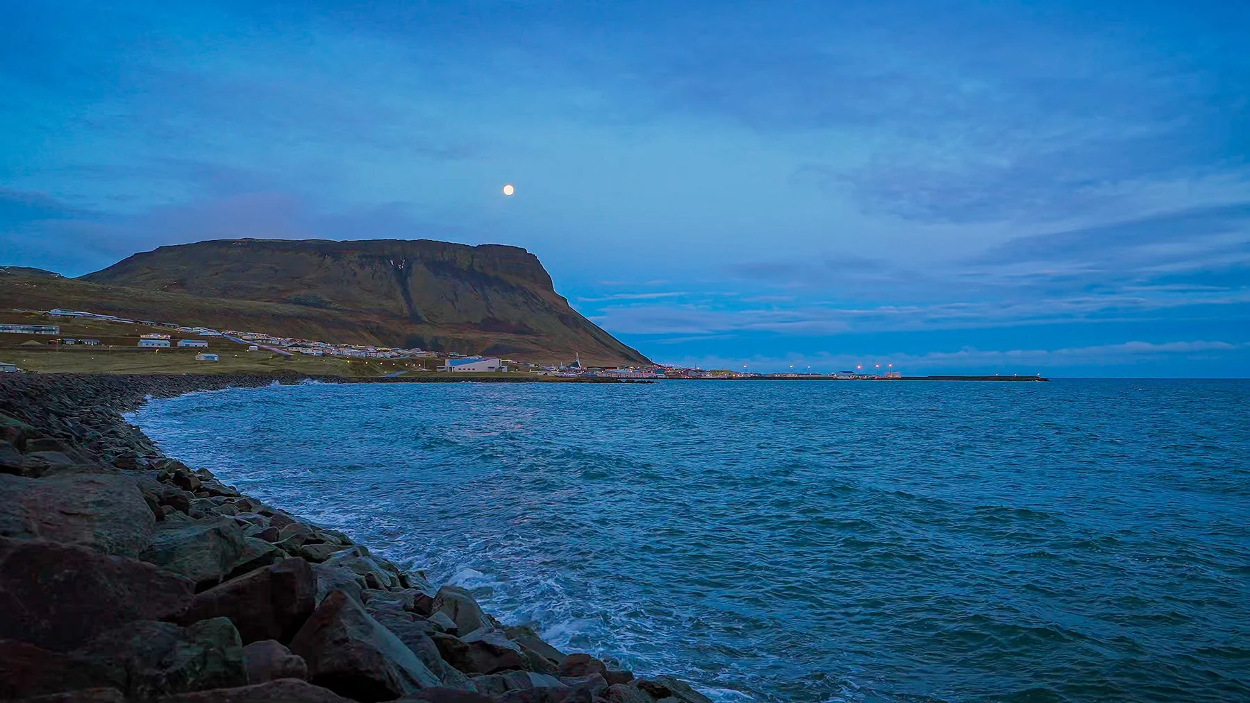 Moonlit Iceland coast at blue hour with calm ocean water, distant shoreline, soft evening light, and a peaceful coastal mood.