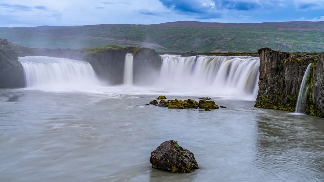 Wide Iceland waterfall flowing through a rocky canyon with a pale glacial river, mossy edges, and open northern landscape.