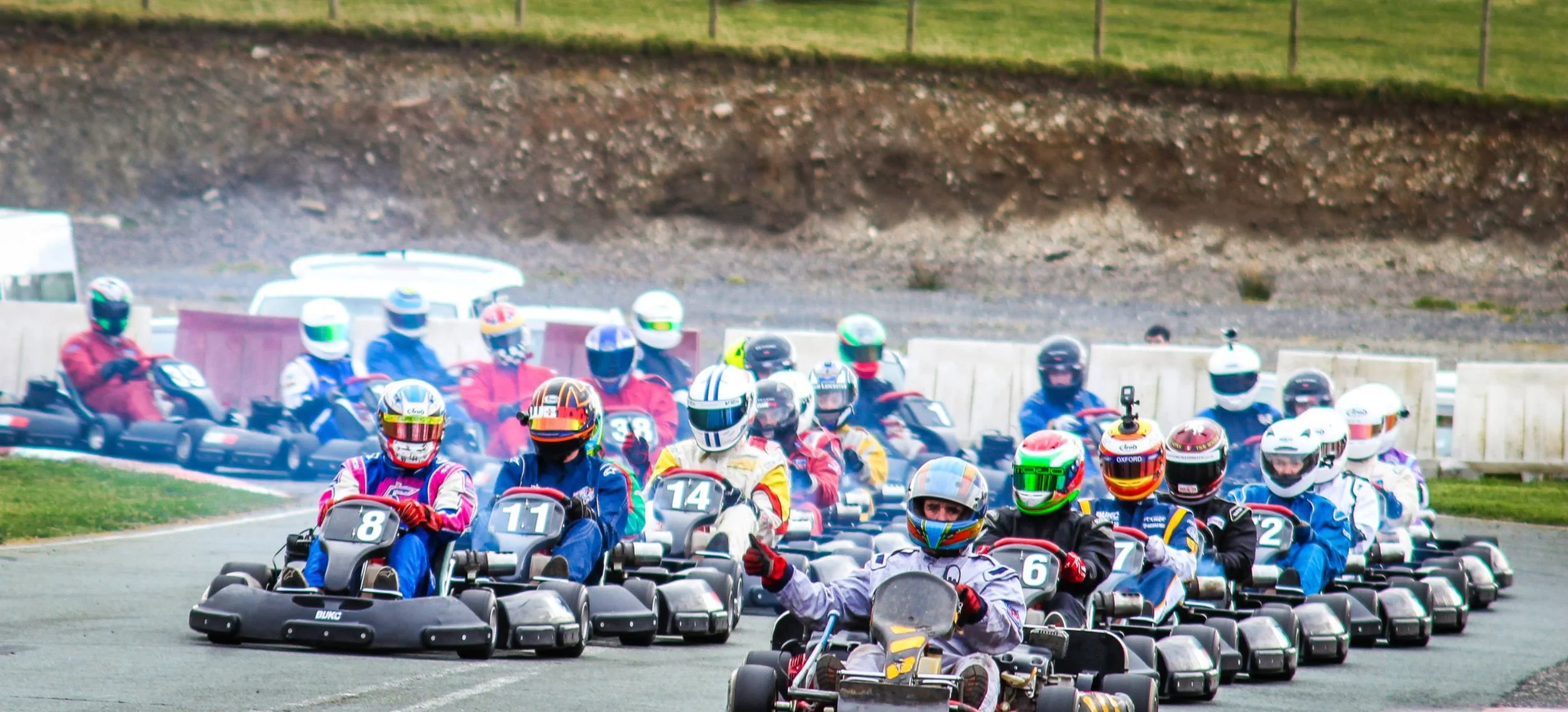 A group of go-kart racers wearing helmets and racing suits on a track, ready to race.