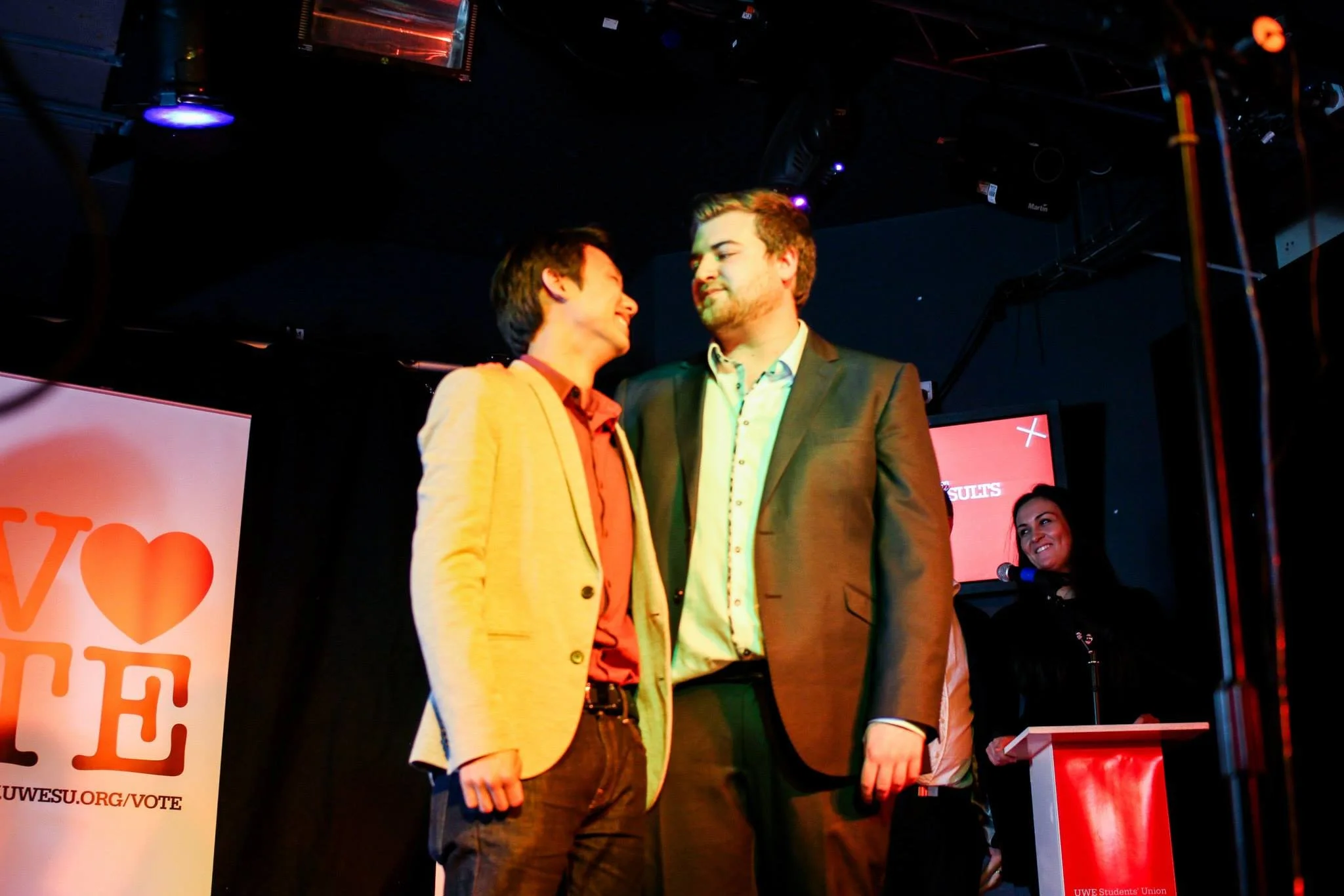 Two men in suits standing closely and smiling at each other on stage during a rally, with a woman at a podium and a UWE Students' Union banner nearby.