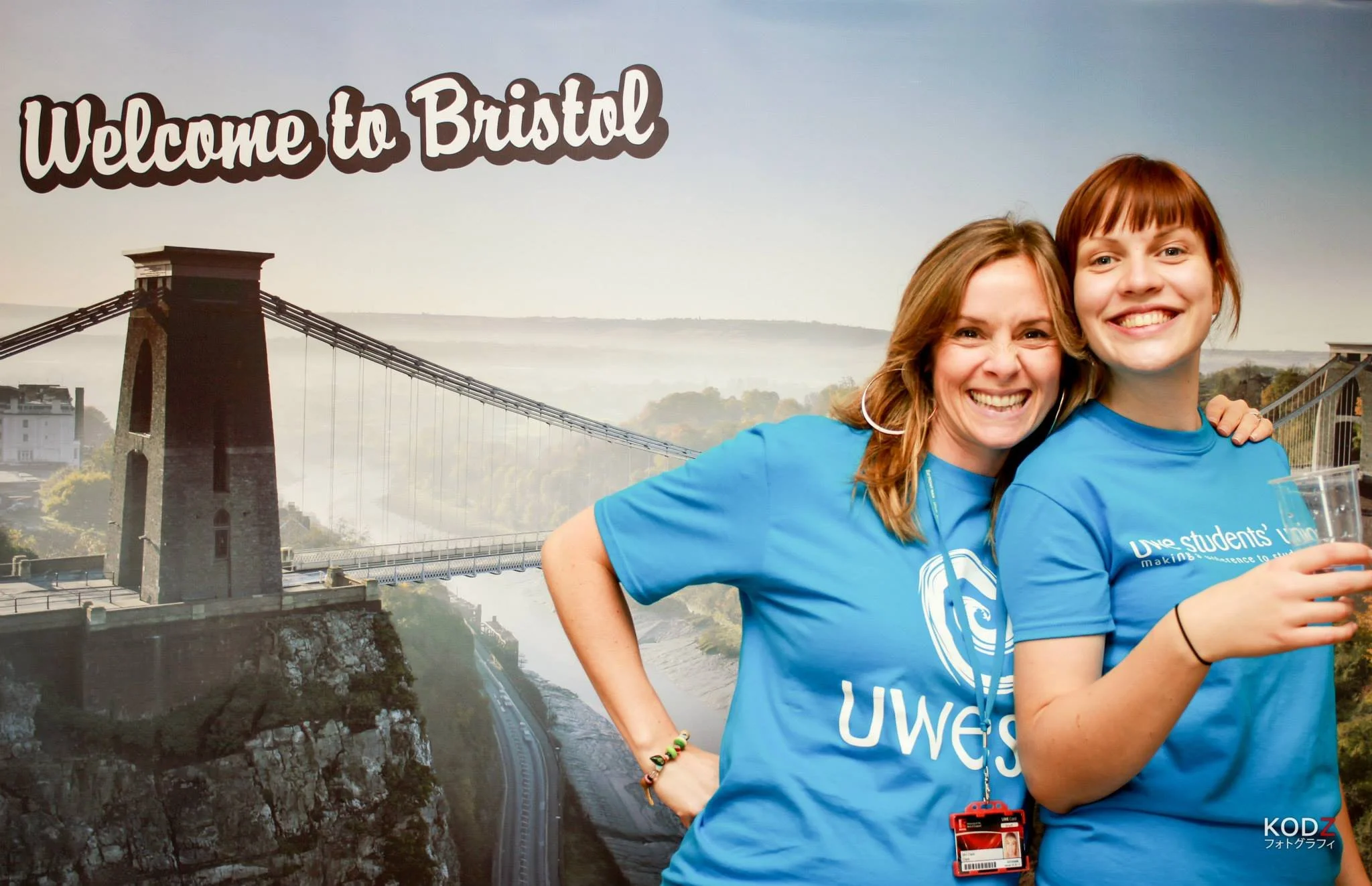 Two smiling women wearing blue UWE shirts, standing in front of a large backdrop of the Clifton Suspension Bridge in Bristol, smiling at the camera.