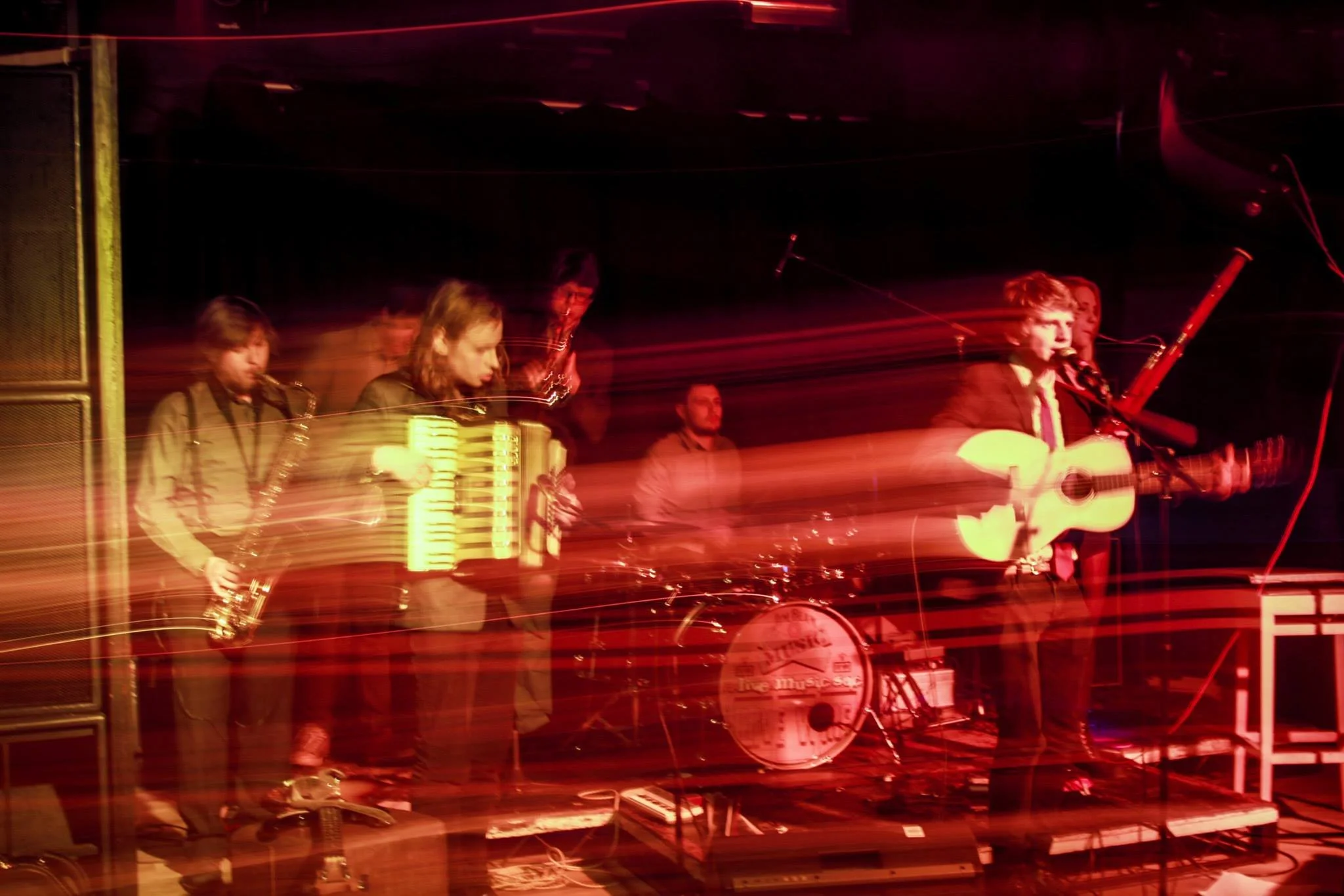 A band performing on stage with colorful light streaks, including members playing saxophone, accordion, drums, and guitar.