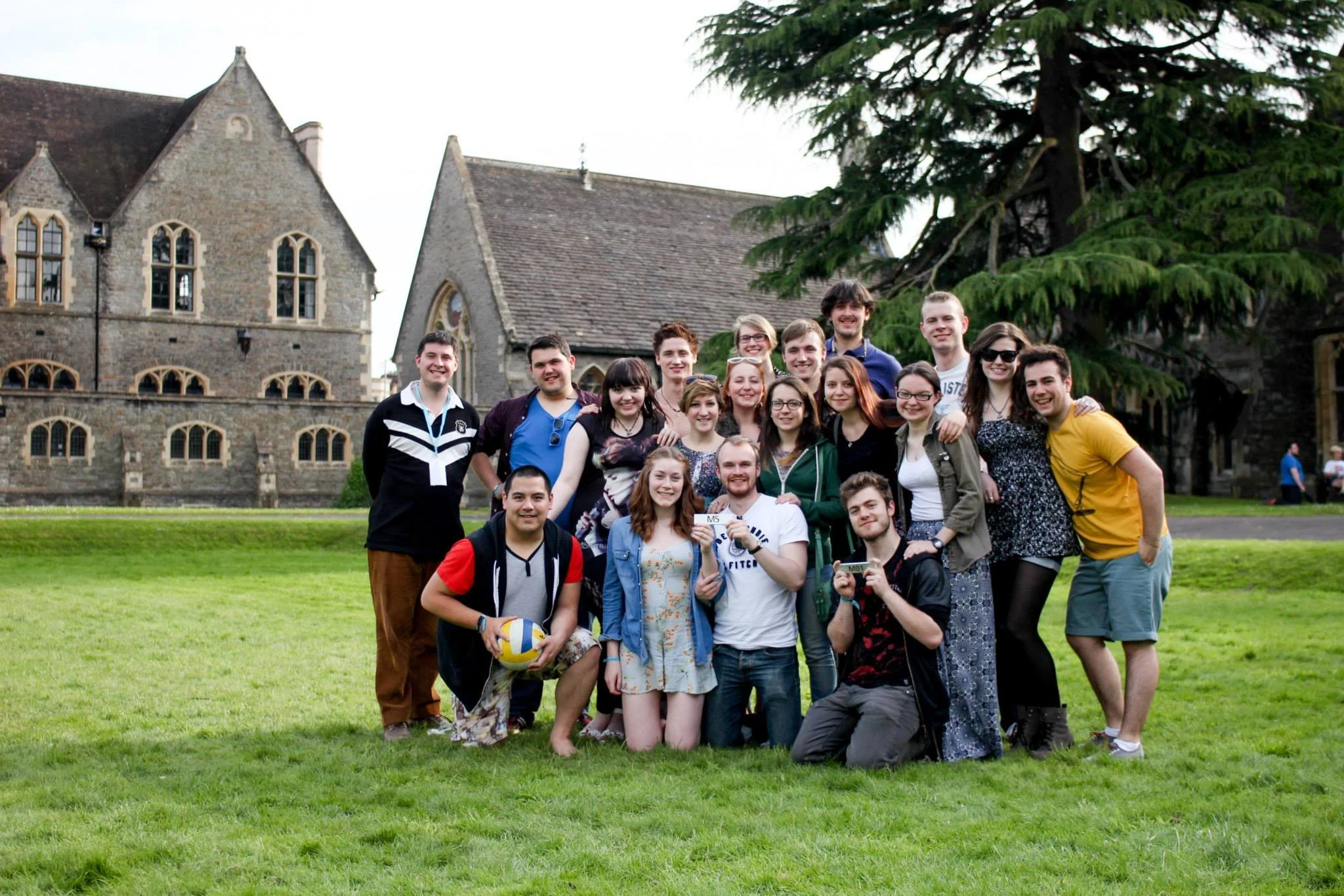 Group of young adults posing outdoors on a grassy lawn in front of a historic stone building with large trees, some holding food and drinks, smiling for the camera.