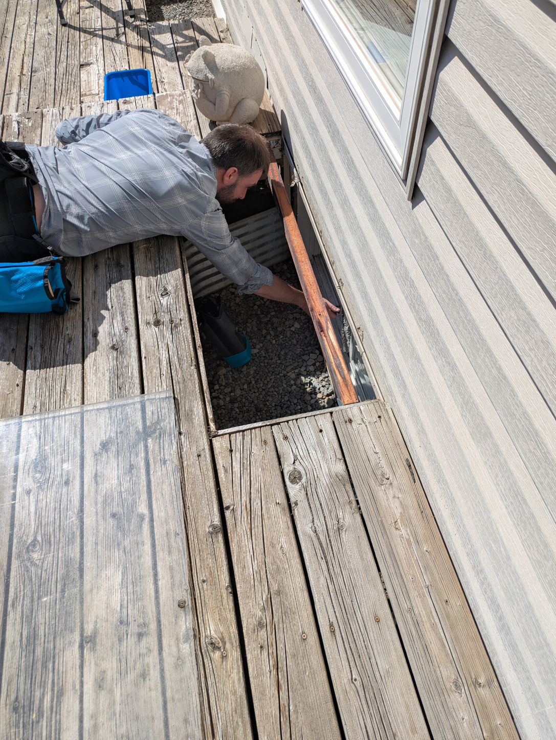 Man inspecting or cleaning an uncovered area of a wooden deck outside a house, with a decorative frog statue and various tools nearby.