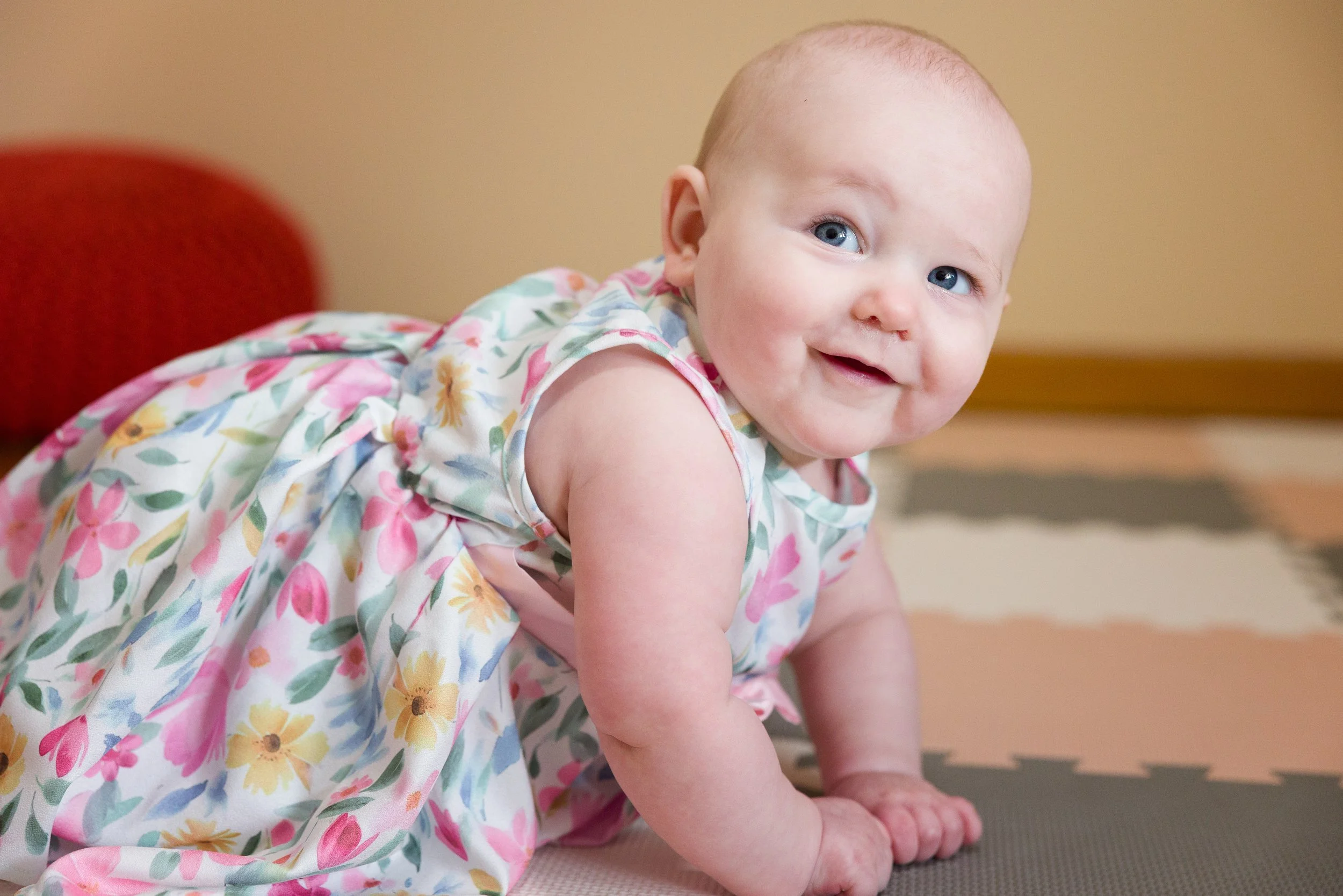 Seven-month-old baby girl in a floral dress smiling during an in-home family photography session.