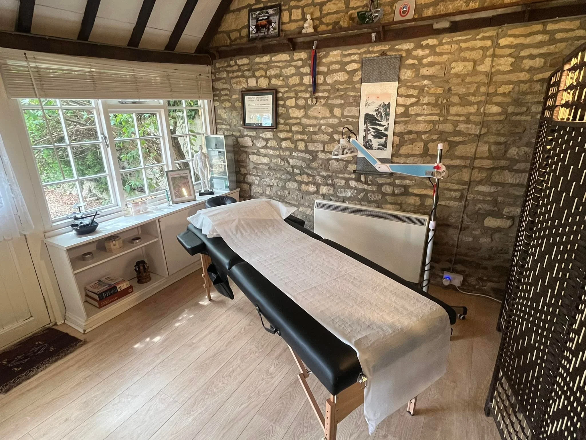 A massage therapy room with a massage table covered with a white sheet, positioned near a window with bamboo blinds. The room has a brick wall decorated with art, a framed certificate, and a mounted light. There are various decorative items on shelves and a small cabinet with books and a sculpture. An adjustable lamp is positioned near the massage table, and a room divider is visible on the right side.