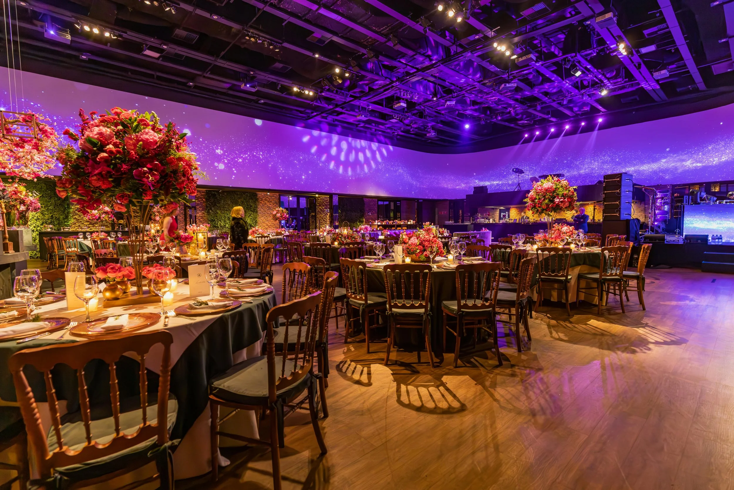 Elegantly decorated banquet hall with round tables covered in black tablecloths, adorned with pink and purple floral centerpieces, set for a formal event, with purple lighting and a large digital starry sky projection on the wall.