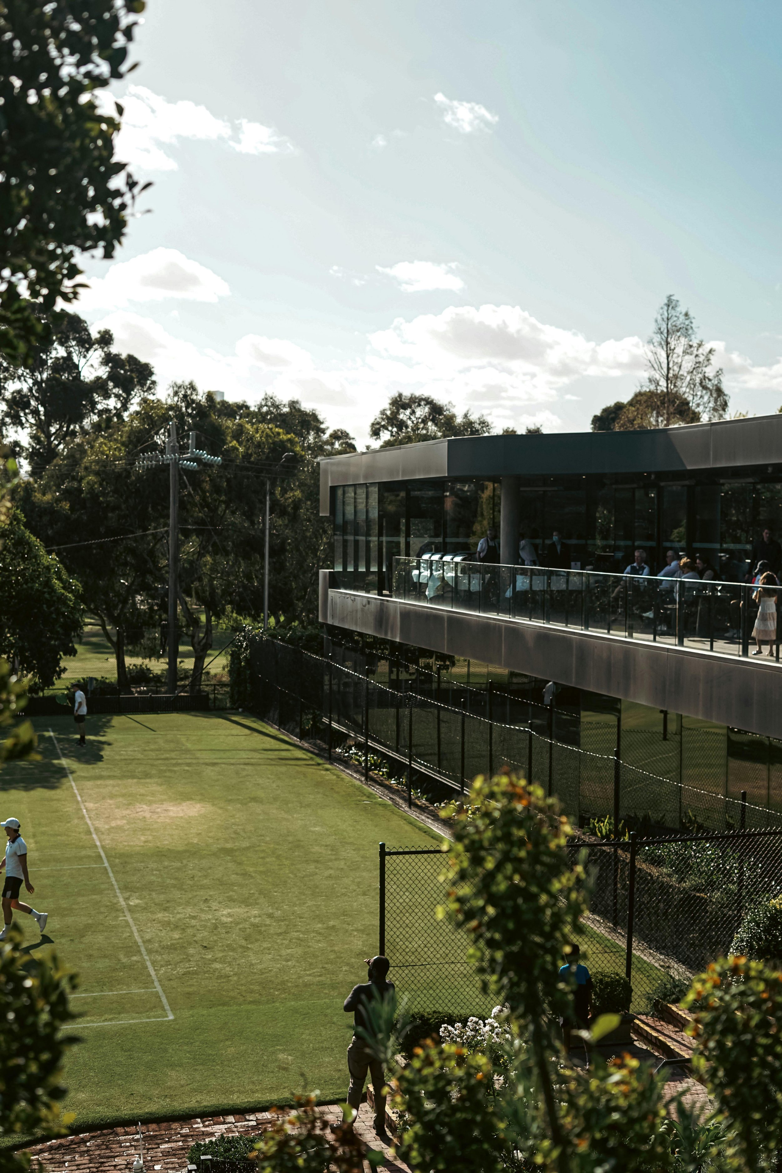 People playing tennis on a grass court outside a modern glass-clad building with a balcony, surrounded by trees and a clear sky.