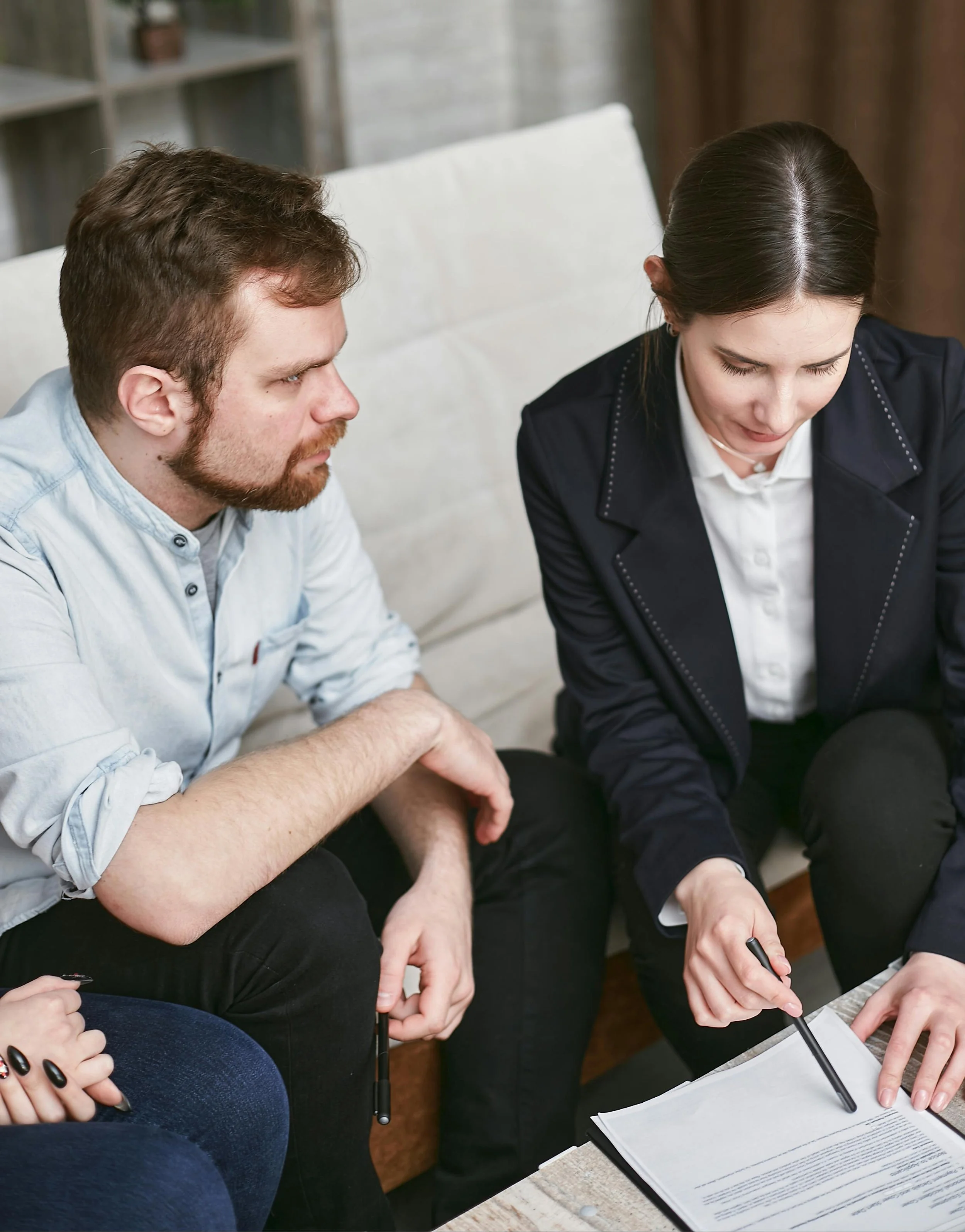 A woman in a dark blazer and white shirt explaining a document to a man with a beard and light shirt in a casual setting.