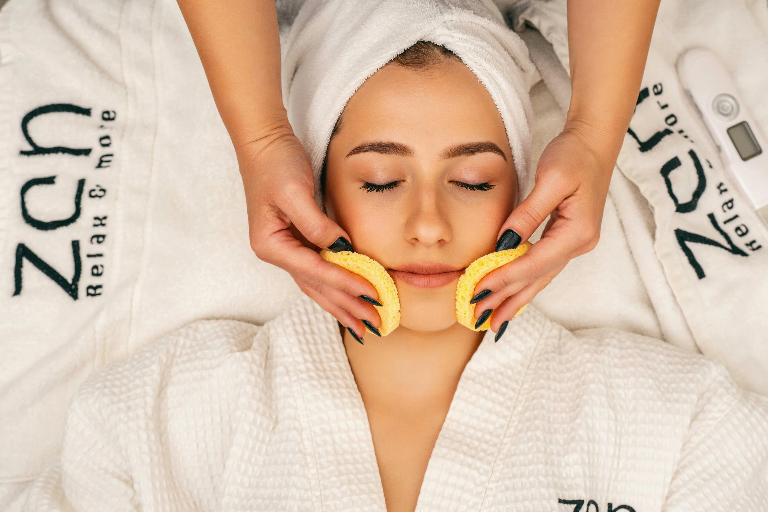 A woman receiving a facial treatment, with her eyes closed, lying on a bed covered with a towel that says 'ZC', in a spa or wellness center.