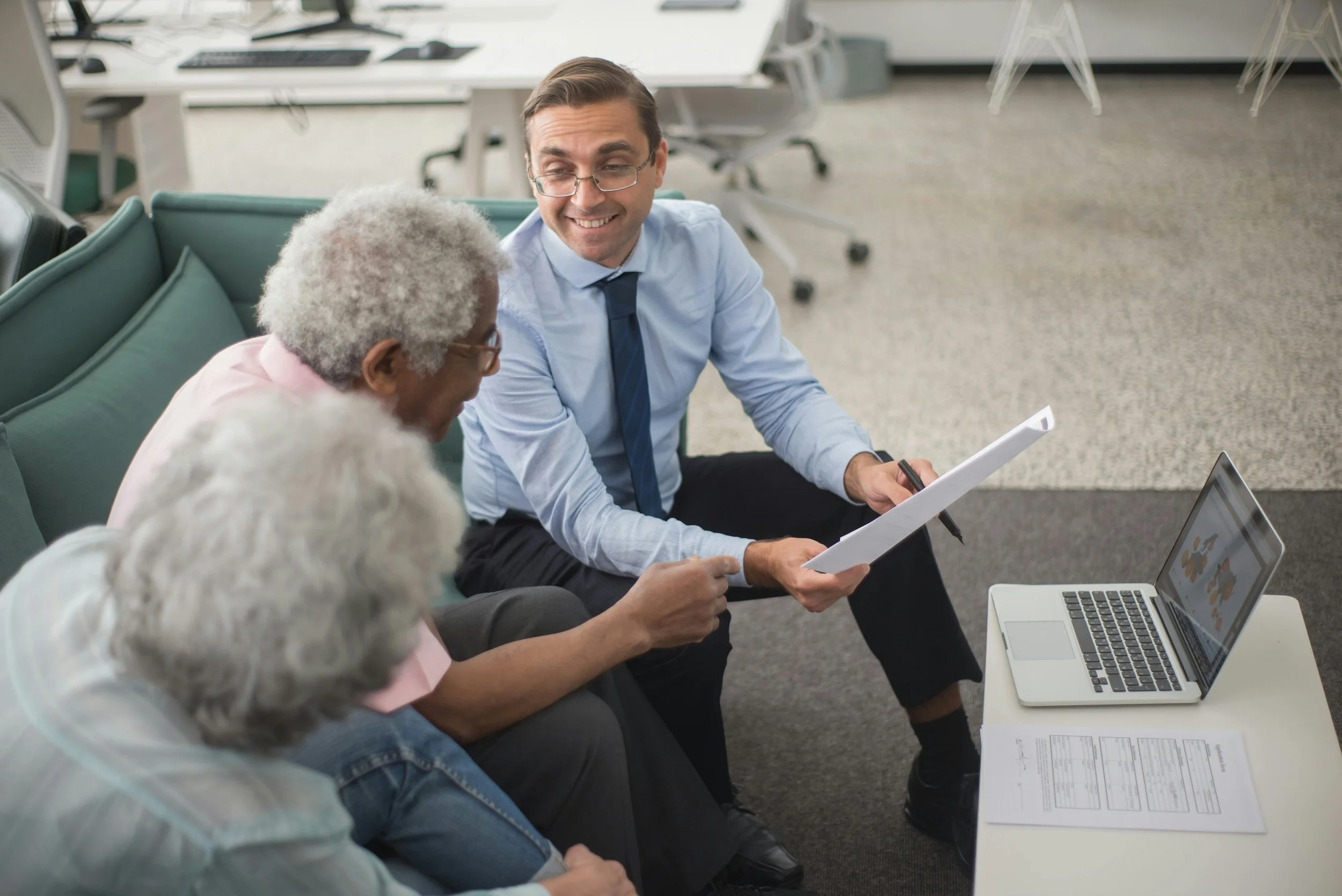 Business meeting with three people discussing documents in an office setting, with a laptop and papers on a table.