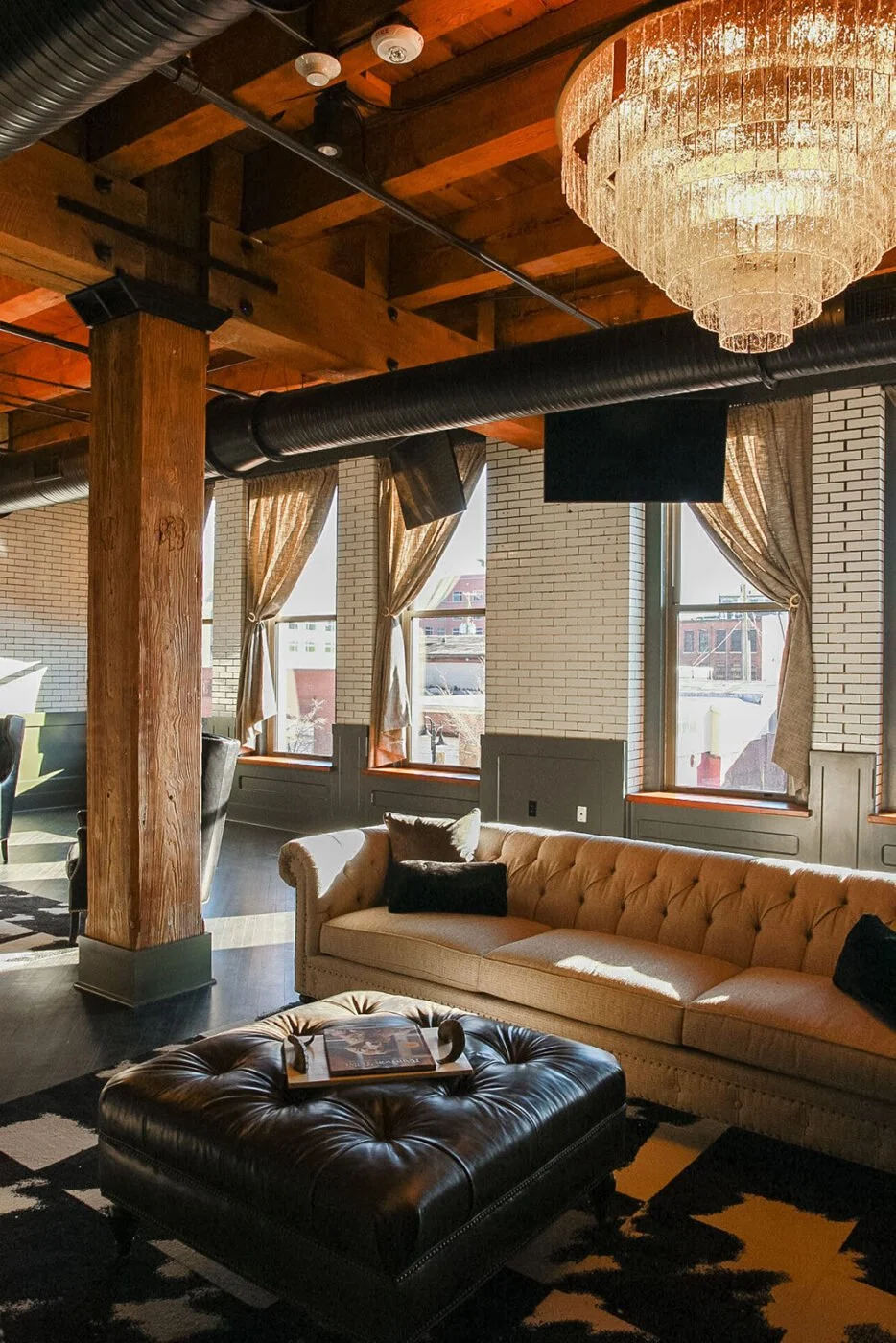 A cozy lounge area with a beige tufted sofa, black throw pillows, and a black leather ottoman with a tray on top. The room has large windows with beige curtains, wooden beams on the ceiling, and a chandelier. Exposed brick walls and black ductwork are visible.