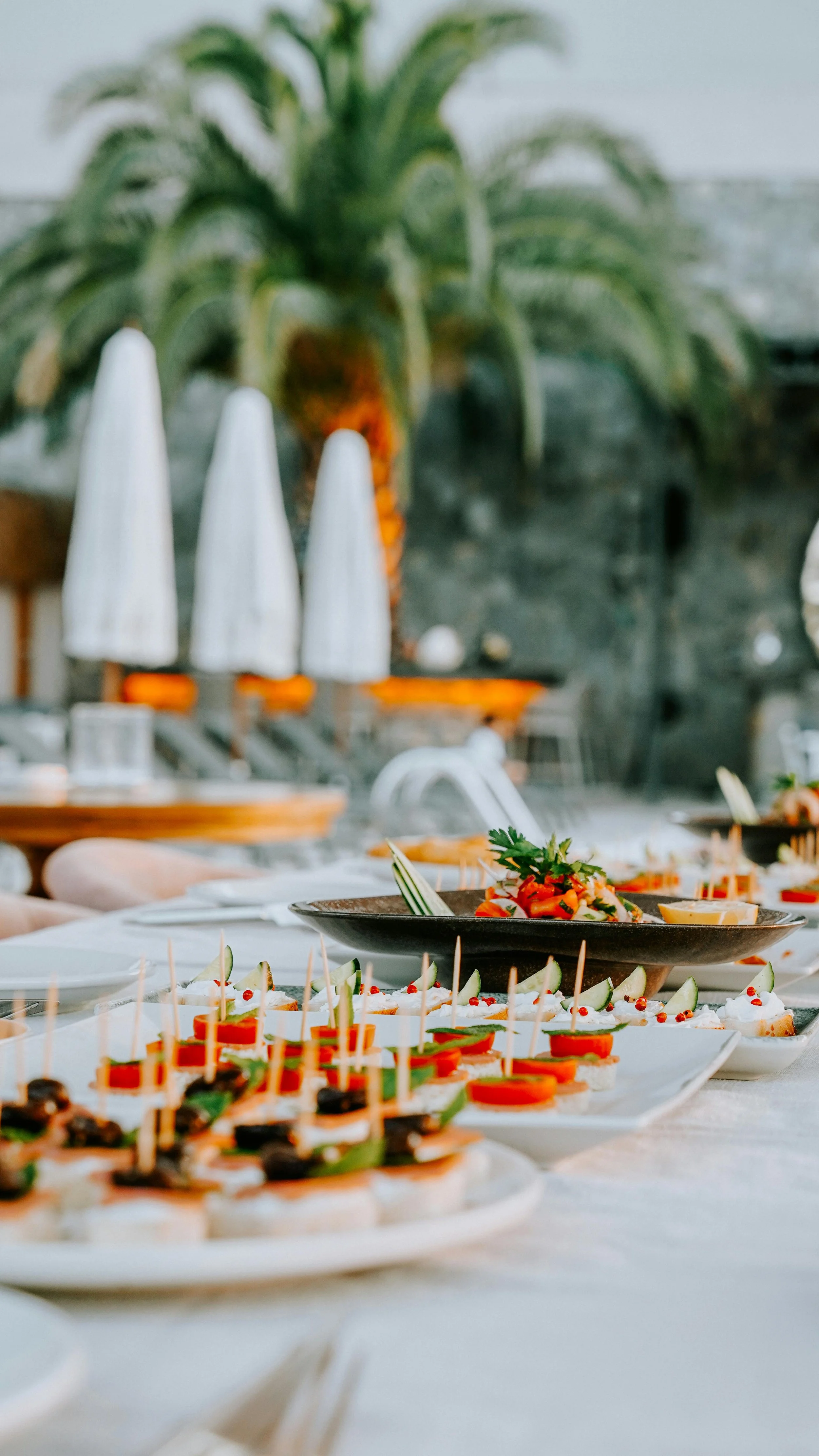 A table set with various appetizers and small dishes, including skewered bites and garnished salads, with a large palm plant and modern decor in the background.