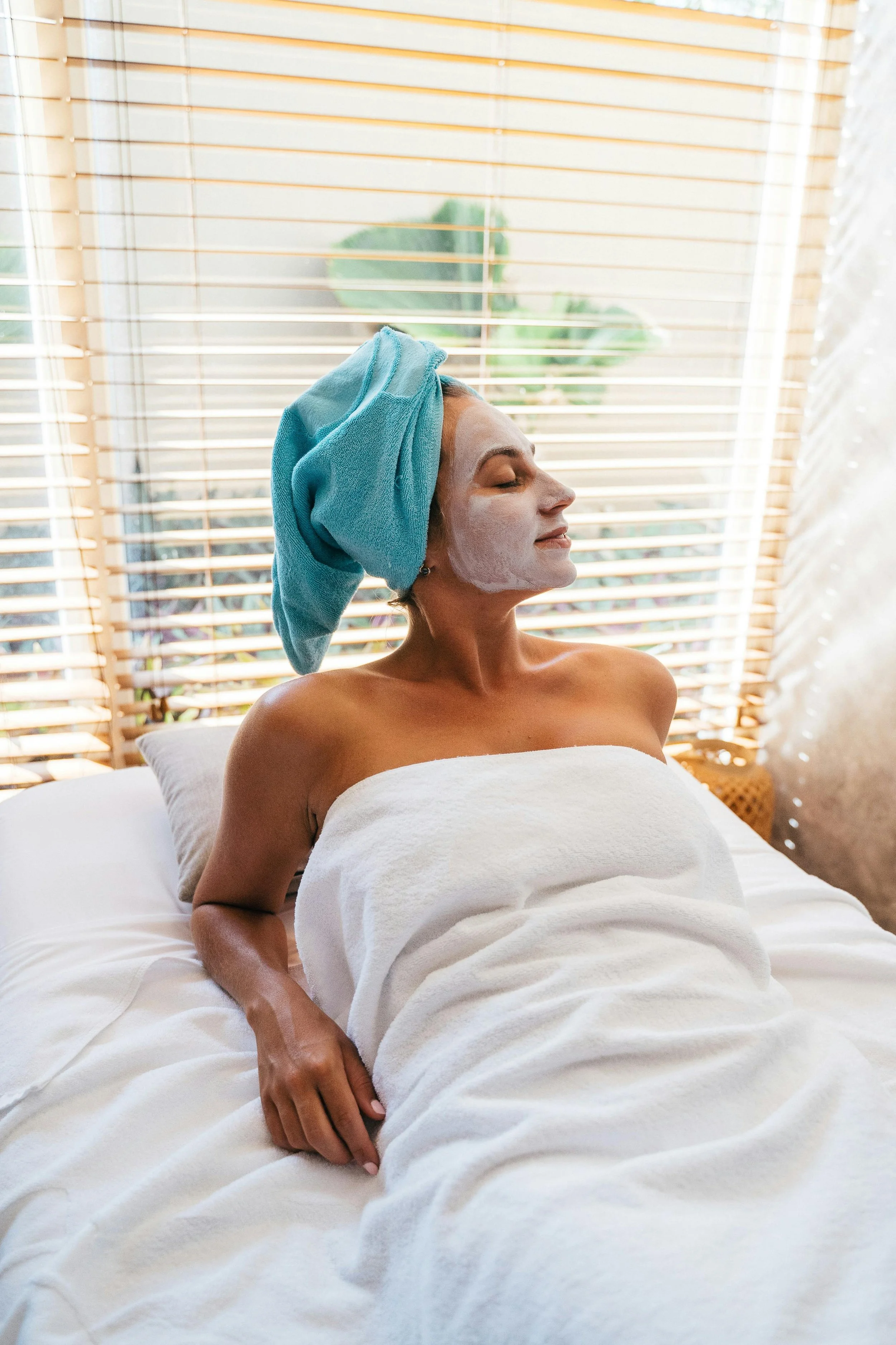 A woman relaxing in a spa with a facial mask, towel wrapped around her body, and a towel wrapped around her head, sitting on a bed near a window with blinds.