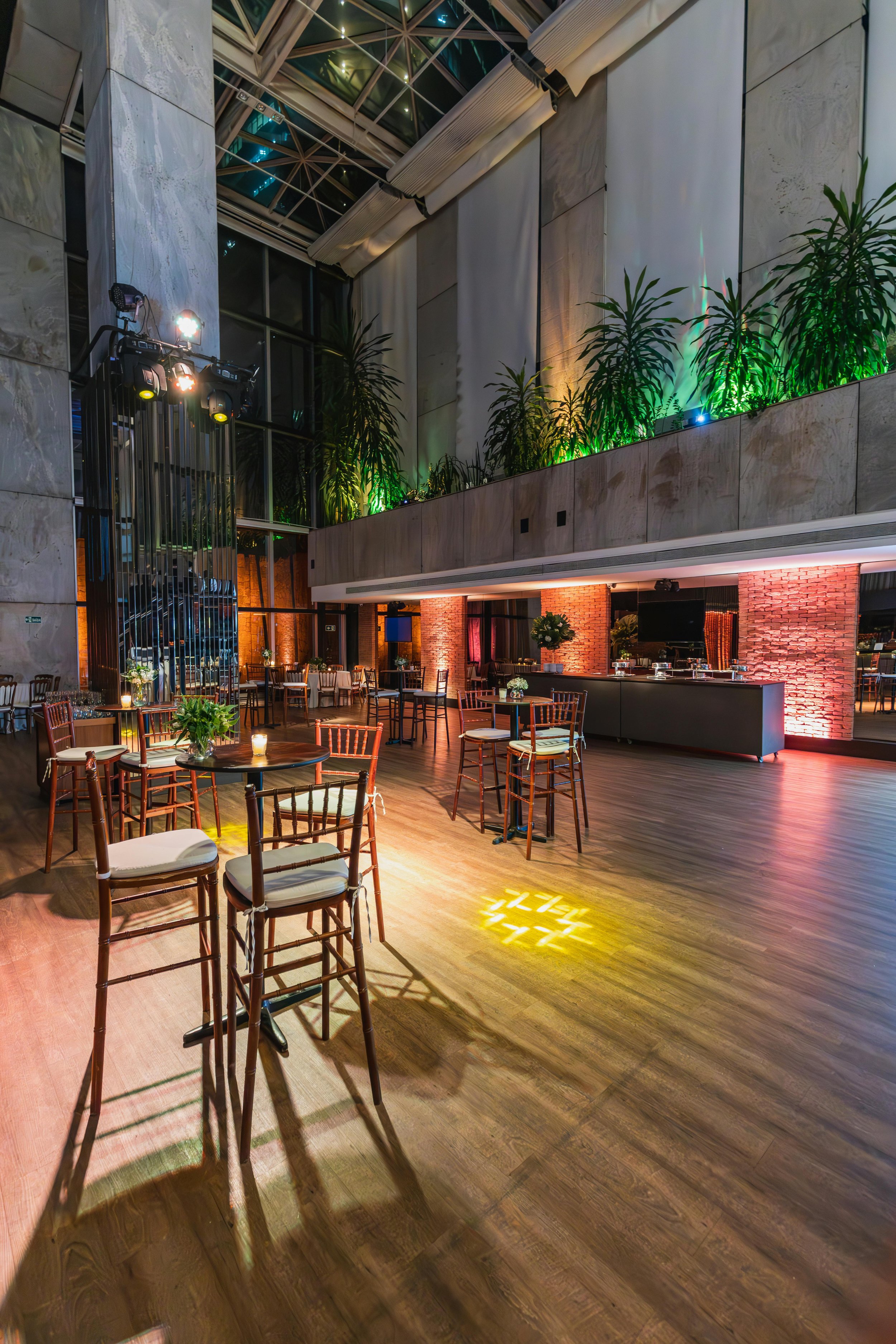 An empty event space or restaurant with high ceilings, wood flooring, tables, chairs, and decorative plants, illuminated by colorful lighting.