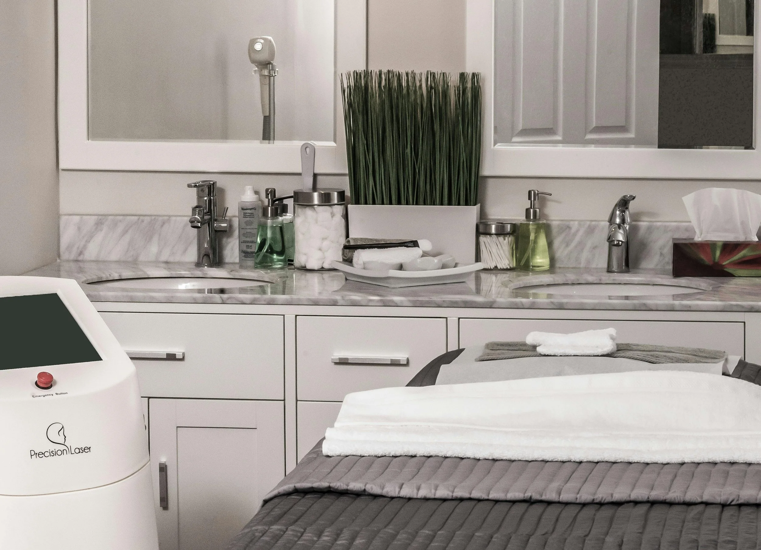 A medical or spa treatment room with a countertop that has various bottles, jars, and a large decorative plant. There are two sinks with silver faucets and a mirror above. Part of a bed with folded towels and a laser device labeled "Precision Laser" is visible in the foreground.