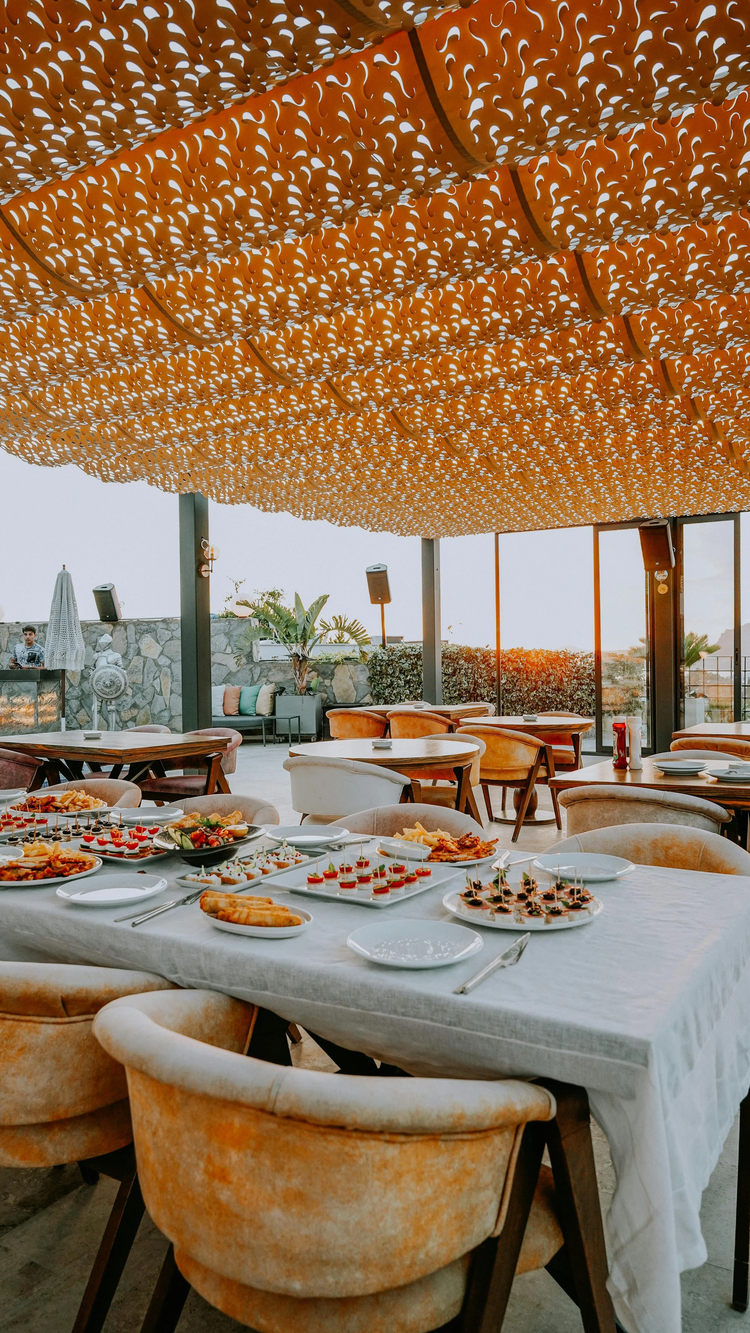 Restaurant or event space with a table set with various appetizers and dishes, decorated with a large orange patterned canopy overhead and a view of a patio with outdoor seating and greenery in the background.