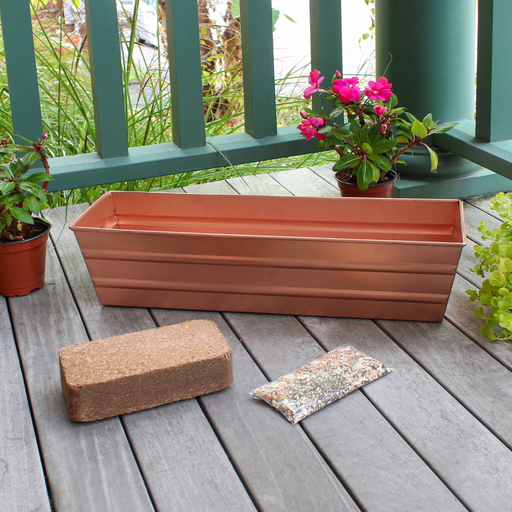 A wooden deck displaying a copper-plated metal window box, a coir brick, a plastic bag of fertilizer, and several potted plants.