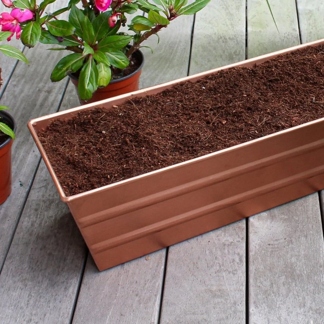 An up-close photo of a rectangular copper-plated window box filled with soil. The windowbox is resting on a wooden deck next to potted plants with pink flowers.