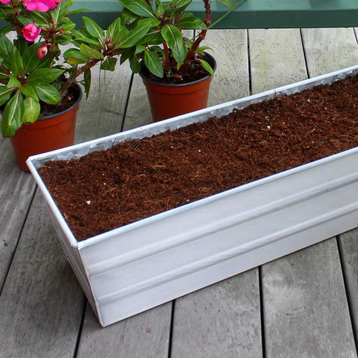 An up-close photo of a rectangular white window box filled with soil. The windowbox is resting on a wooden deck next to potted plants with pink flowers.
