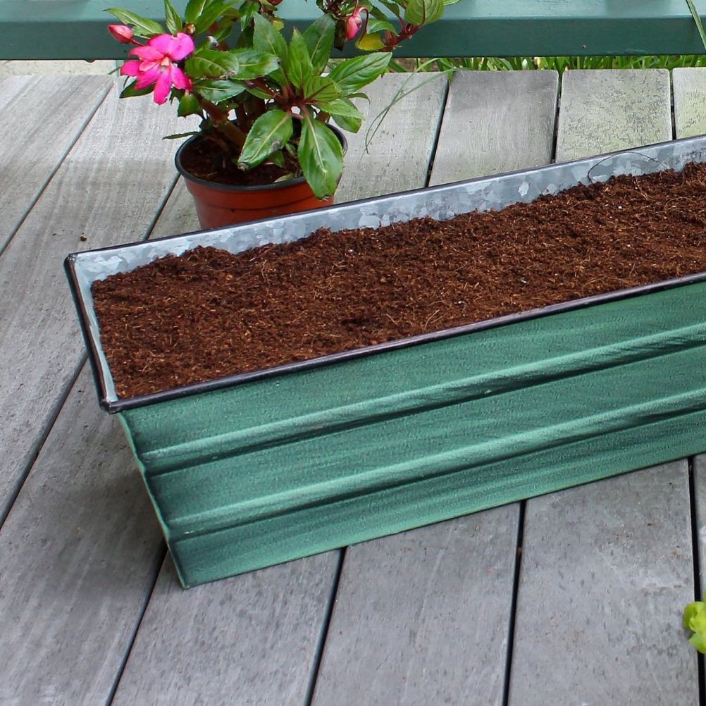 An up-close photo of a rectangular green window box filled with soil. The windowbox is resting on a wooden deck next to potted plants with pink flowers.
