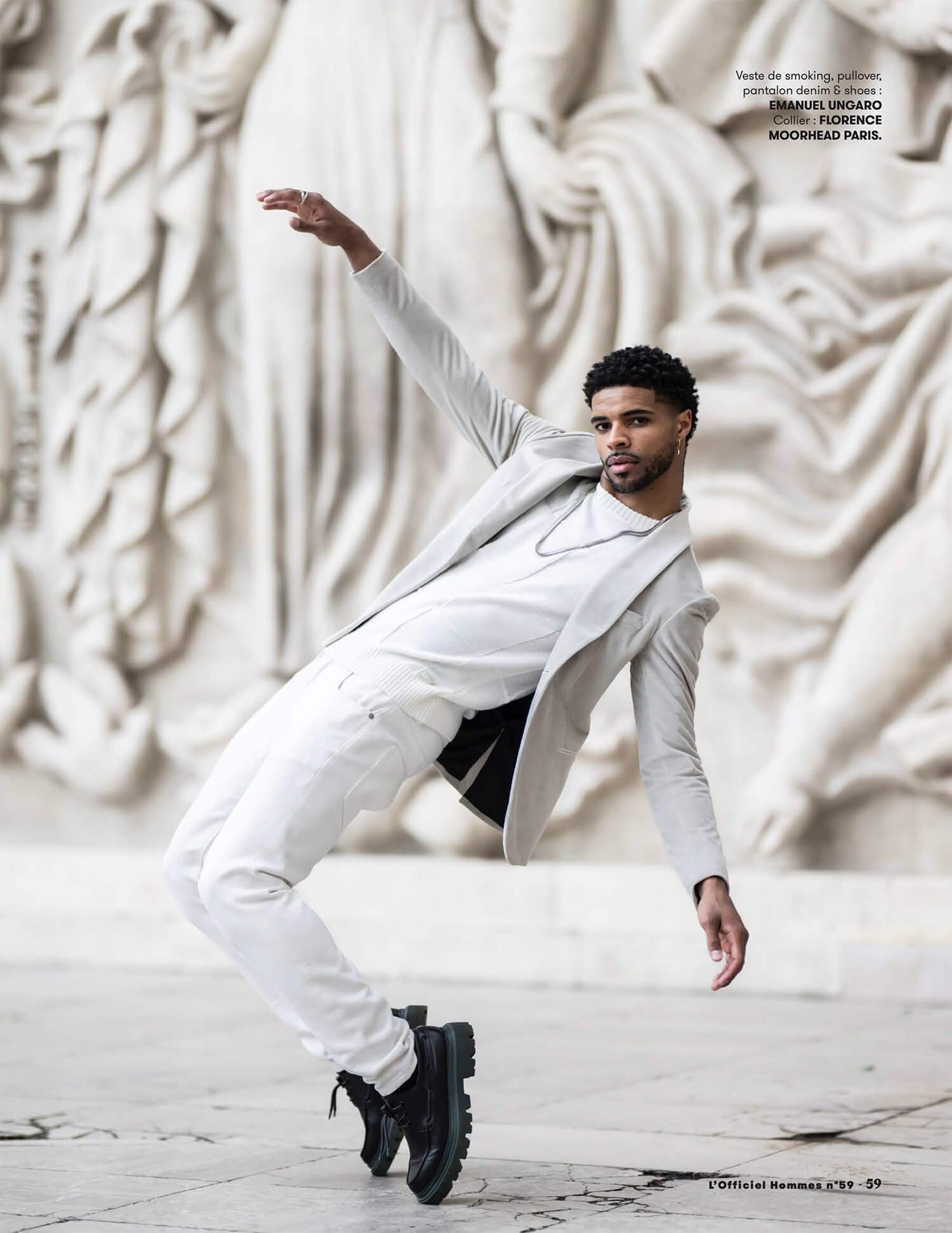 Un homme posant dans une tenue blanche composée d'une veste, pull, pantalon et chaussures noires, devant une sculpture en relief.