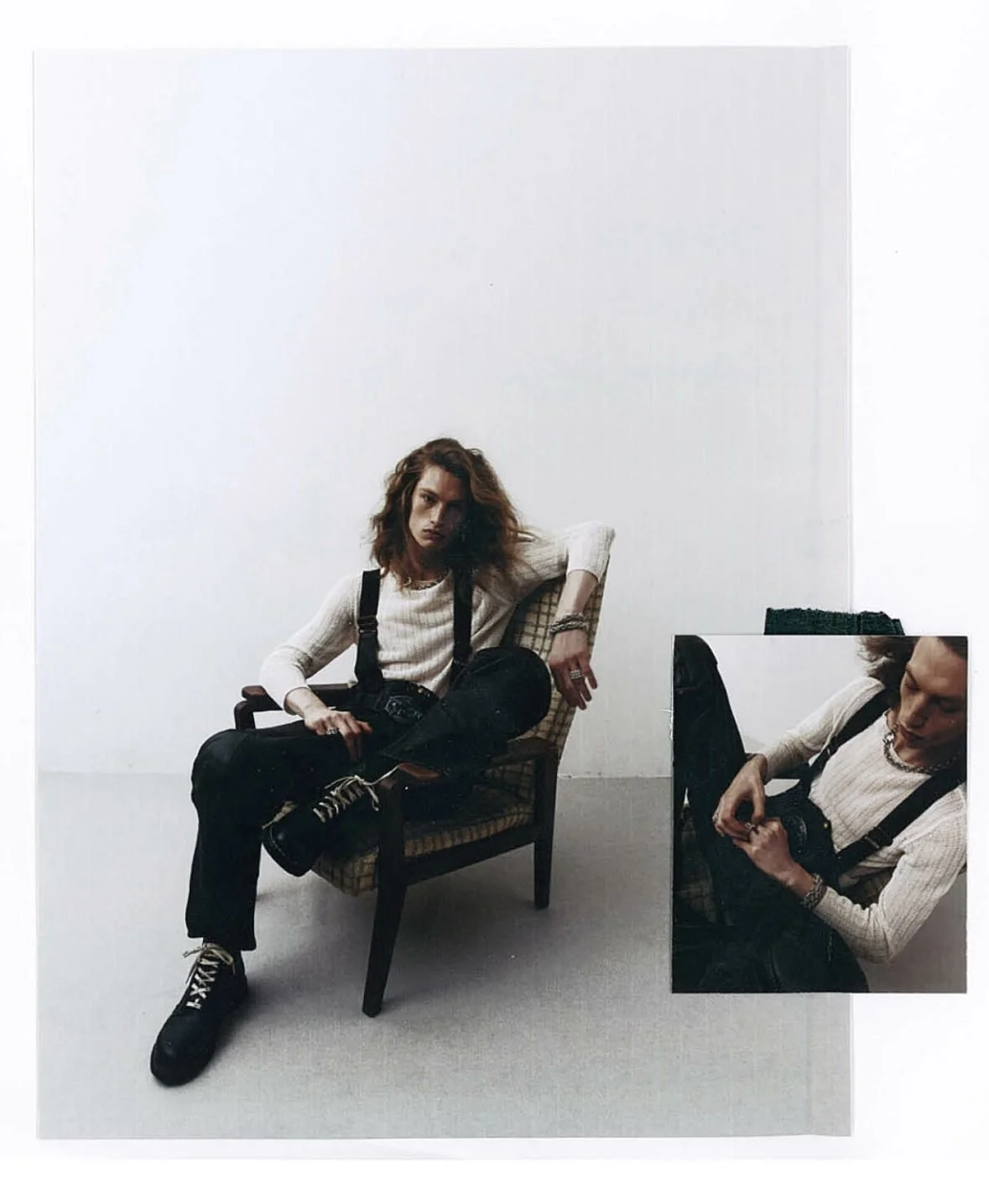 Une homme avec des cheveux bouclés, assise sur une chaise en bois, portant un pull blanc, un pantalon noir, et tenant un appareil photo. Il y a un miroir à côté d'elle qui reflète sa pose.