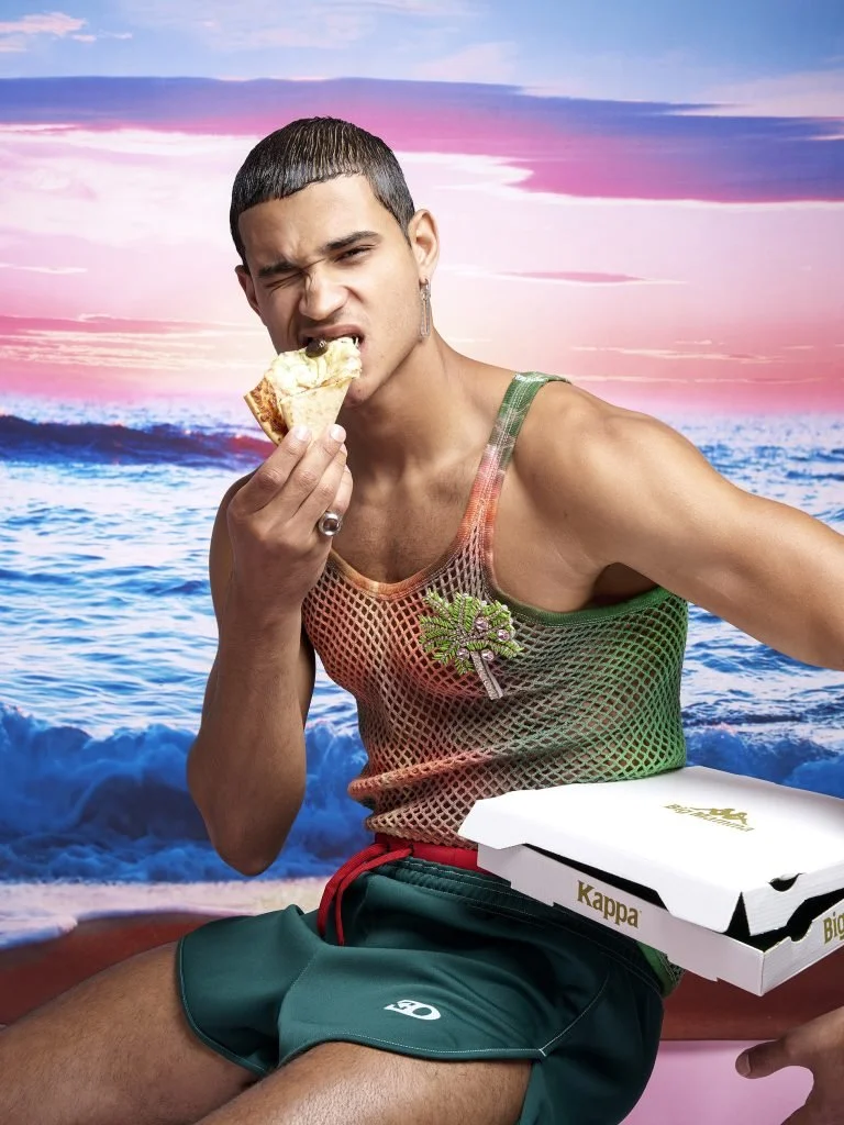 Jeune homme assis sur une plage, mangeant une part de pizza, portant une bague argentée