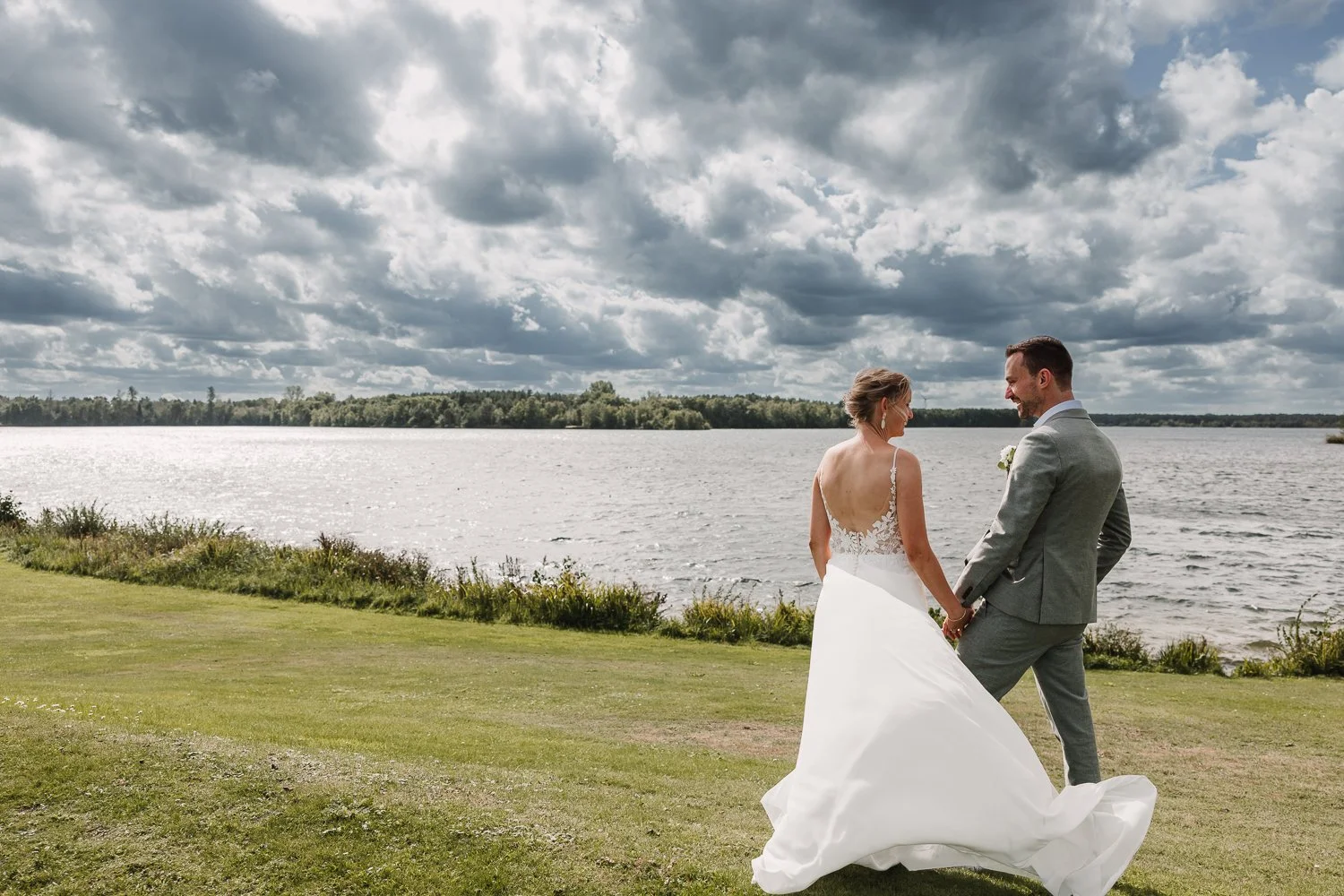 Een bruid en bruidegom die hand in hand staan op een grasveld naast een meer, onder een bewolkte hemel.
