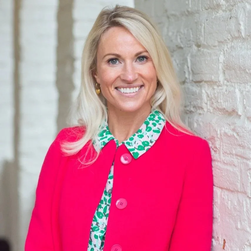 Emily Hill Career Coach smiling at the camera wearing a pink jacket and green shirt leaning against a white wall.