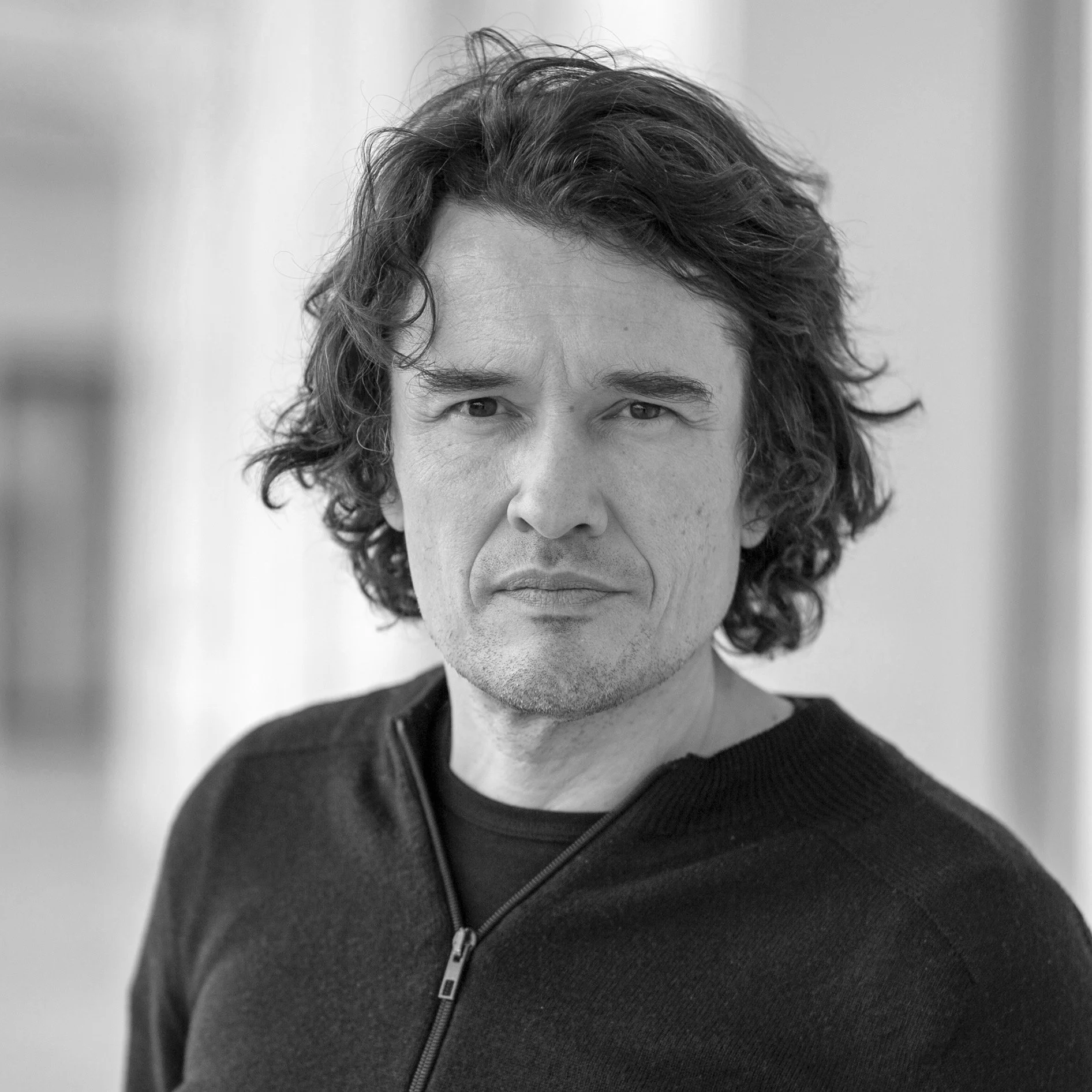 Schwarz-weiß Porträt eines Mannes mit lockigem, mittellangem Haar, der in die Kamera schaut, in einem dunklen Zipper-Top. Der Hintergrund ist unscharf.